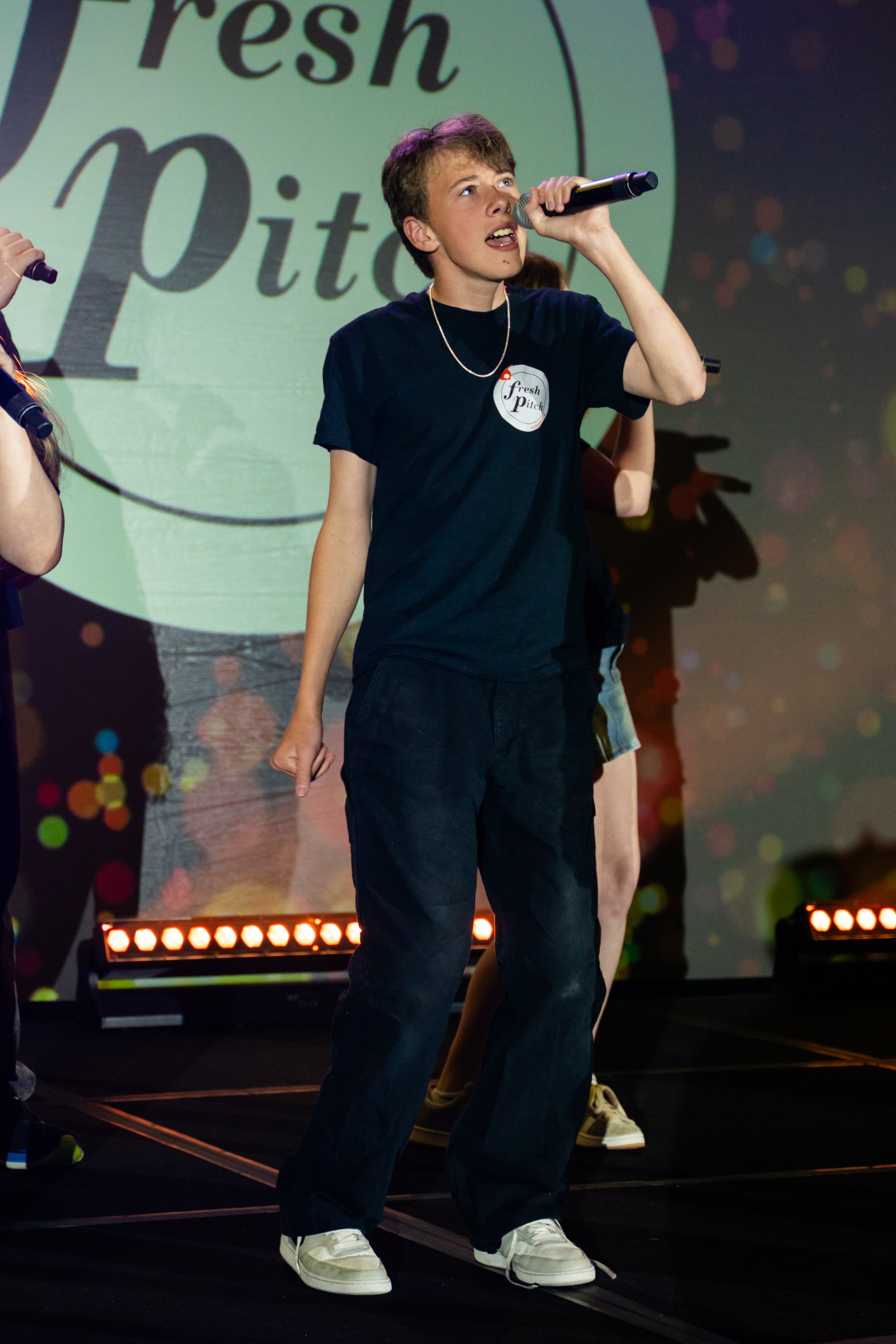 A young man singing into a microphone on a stage with a backdrop that reads 'Fresh Pitch.' He is wearing a black t-shirt, black pants, white sneakers, a gold chain, and has short brown hair. There are stage lights and other performers partially visib