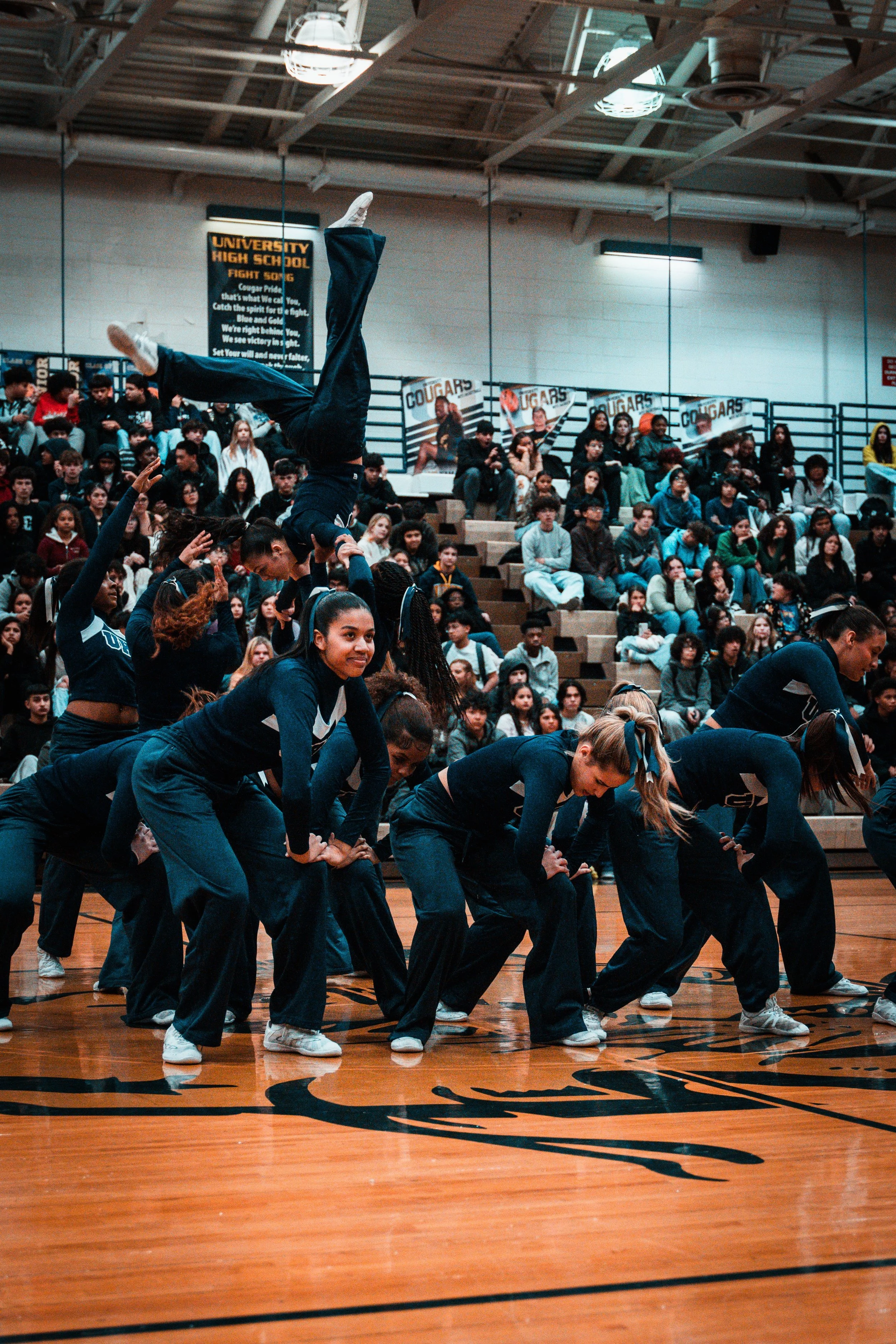 Cheerleaders performing a stunt in a gymnasium during a school event, with audience members seated in bleachers in the background.