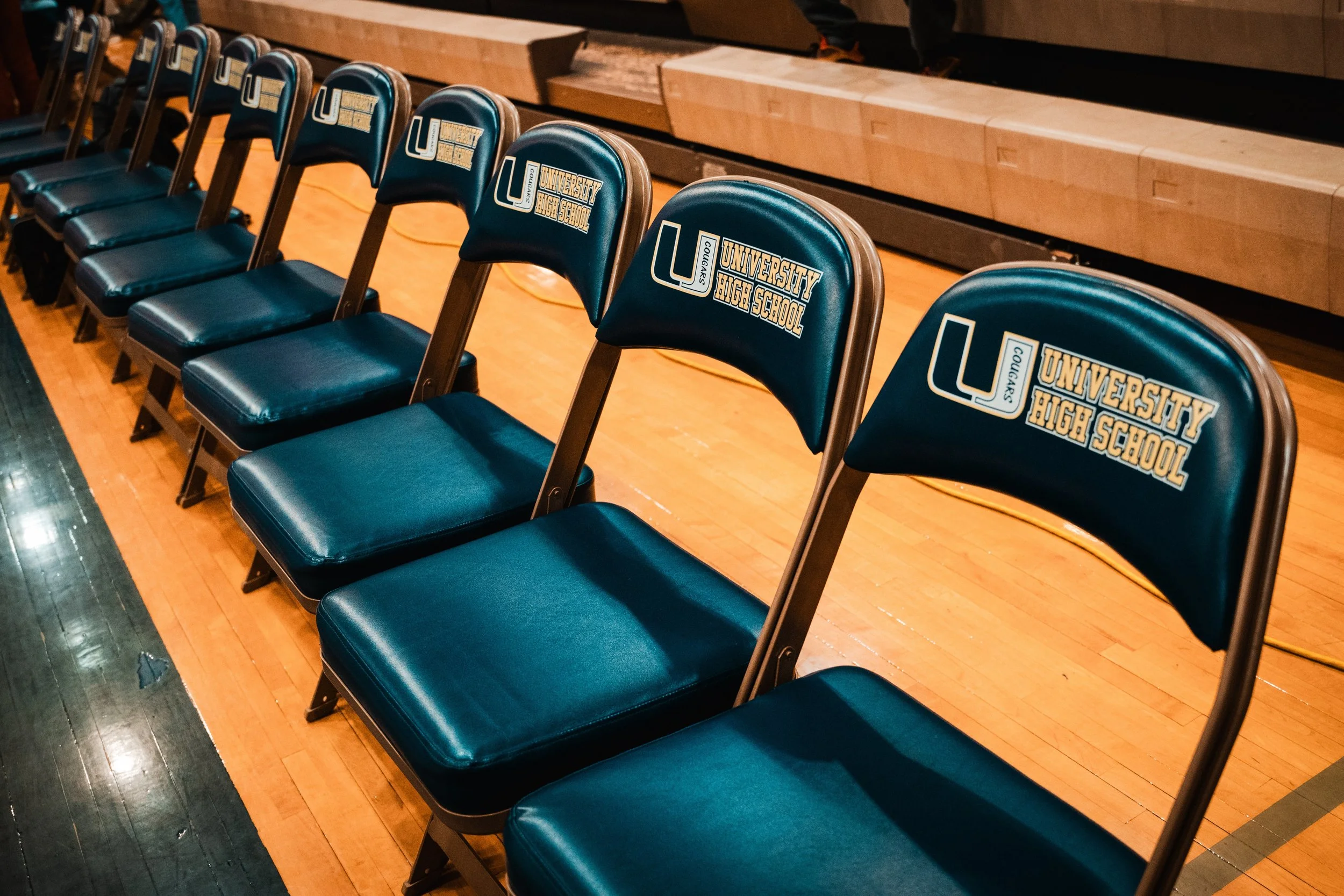 A row of blue chairs with the logo of 'University High School' and the word 'Cougars' on the backrests, placed on a wooden gymnasium floor.