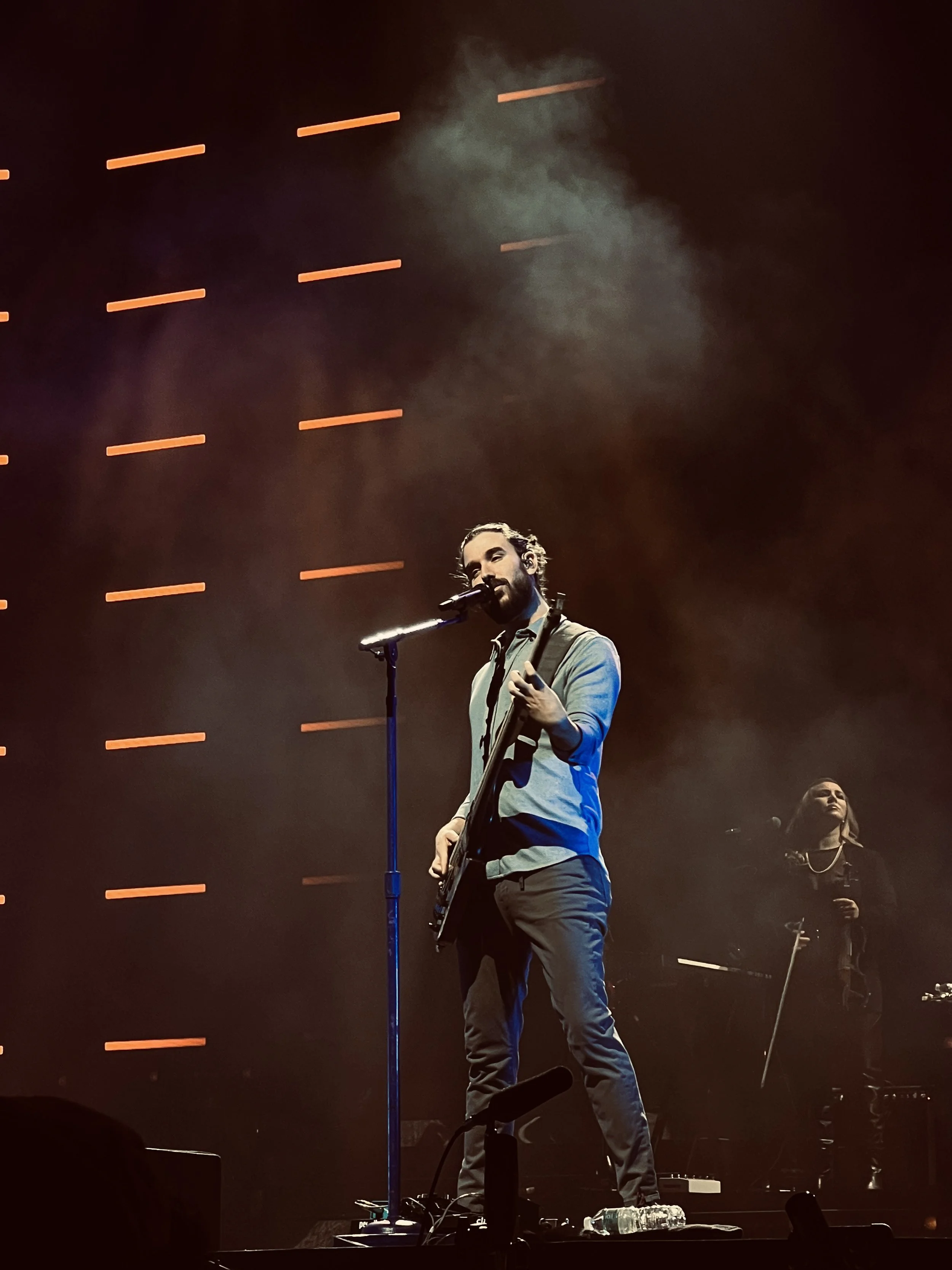 A male musician with long hair and beard, wearing a light gray shirt, performs on stage with a guitar and microphone, while a female backup singer stands behind him. Stage lights create an orange pattern in the background.