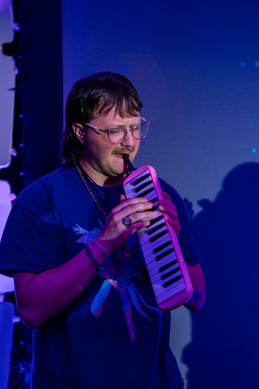 A person with glasses and a necklace playing a melodica in a dimly lit room with purple and blue lighting.