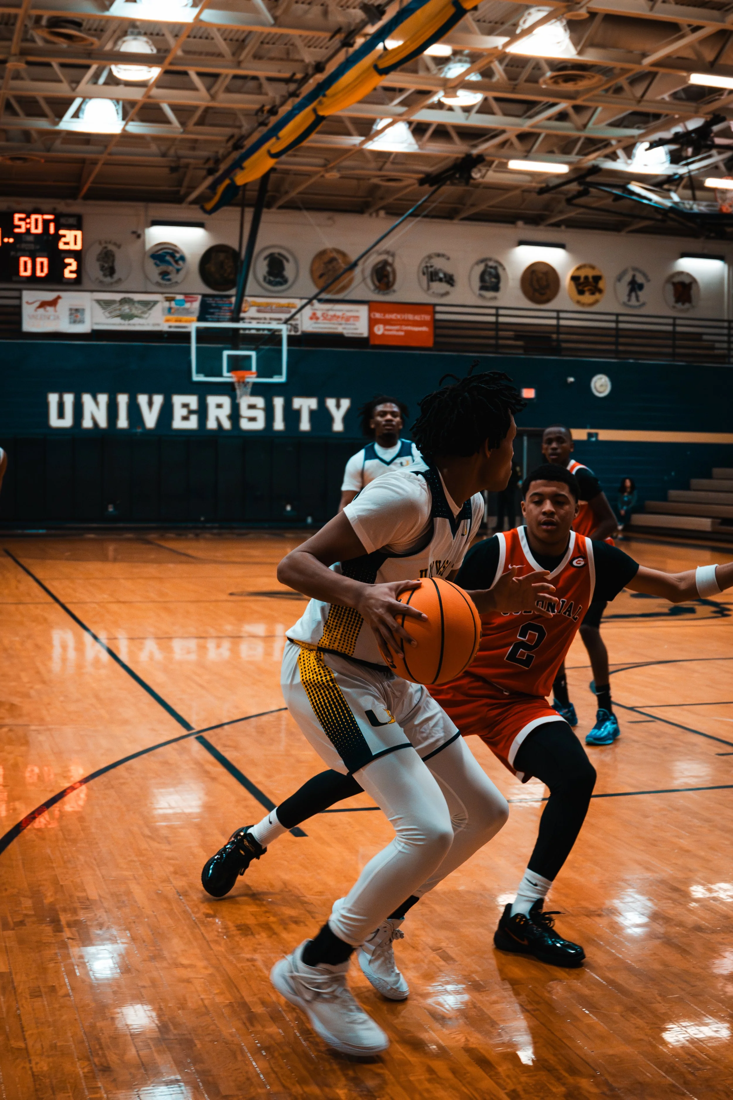 Two basketball players compete on an indoor court, with a scoreboard and banners in the background that say 'UNIVERSITY'.