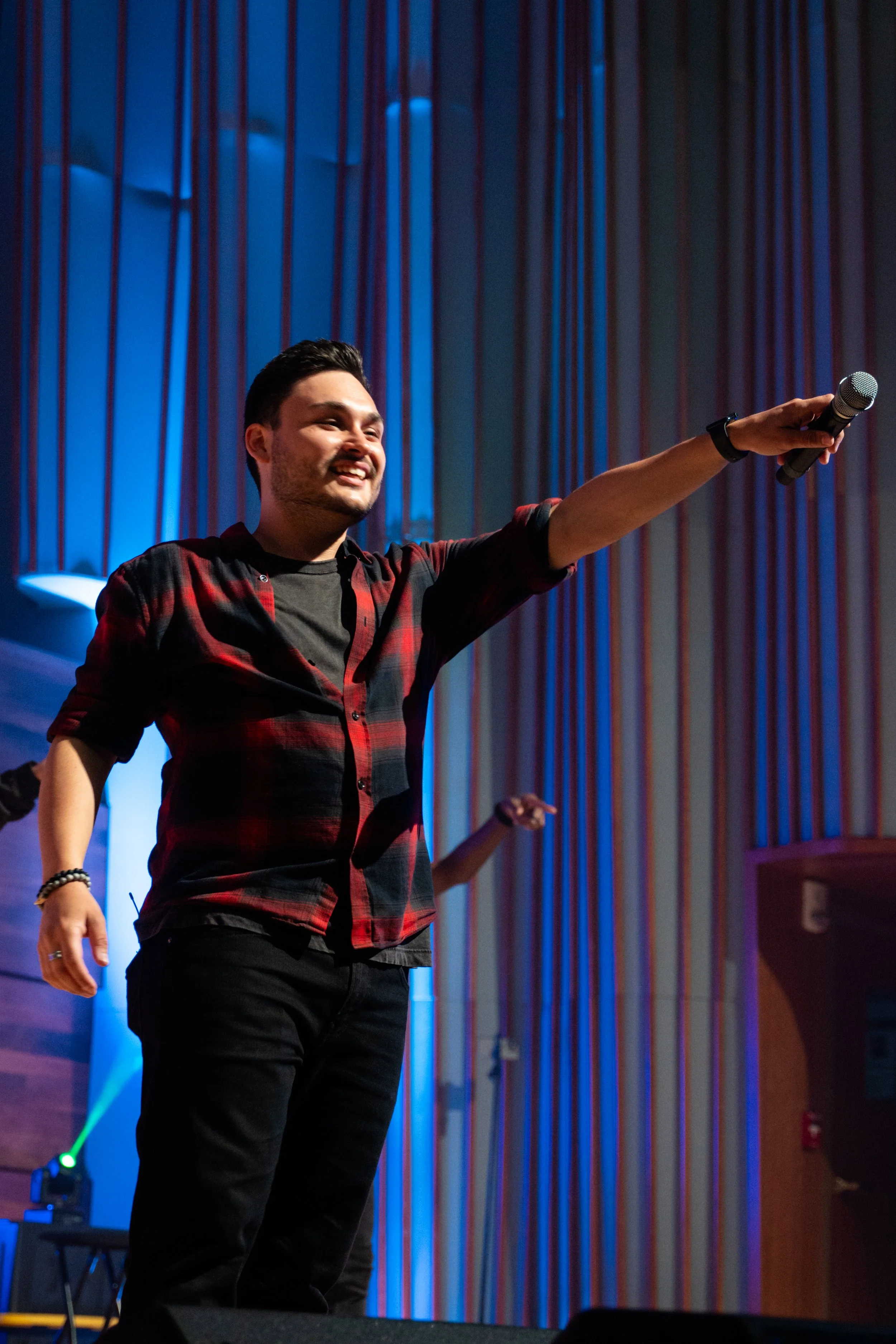 A man with dark hair and a beard, wearing a red plaid shirt over a black t-shirt, smiling and holding a microphone while raising his arm during a performance or presentation on stage with colorful curtains in the background.
