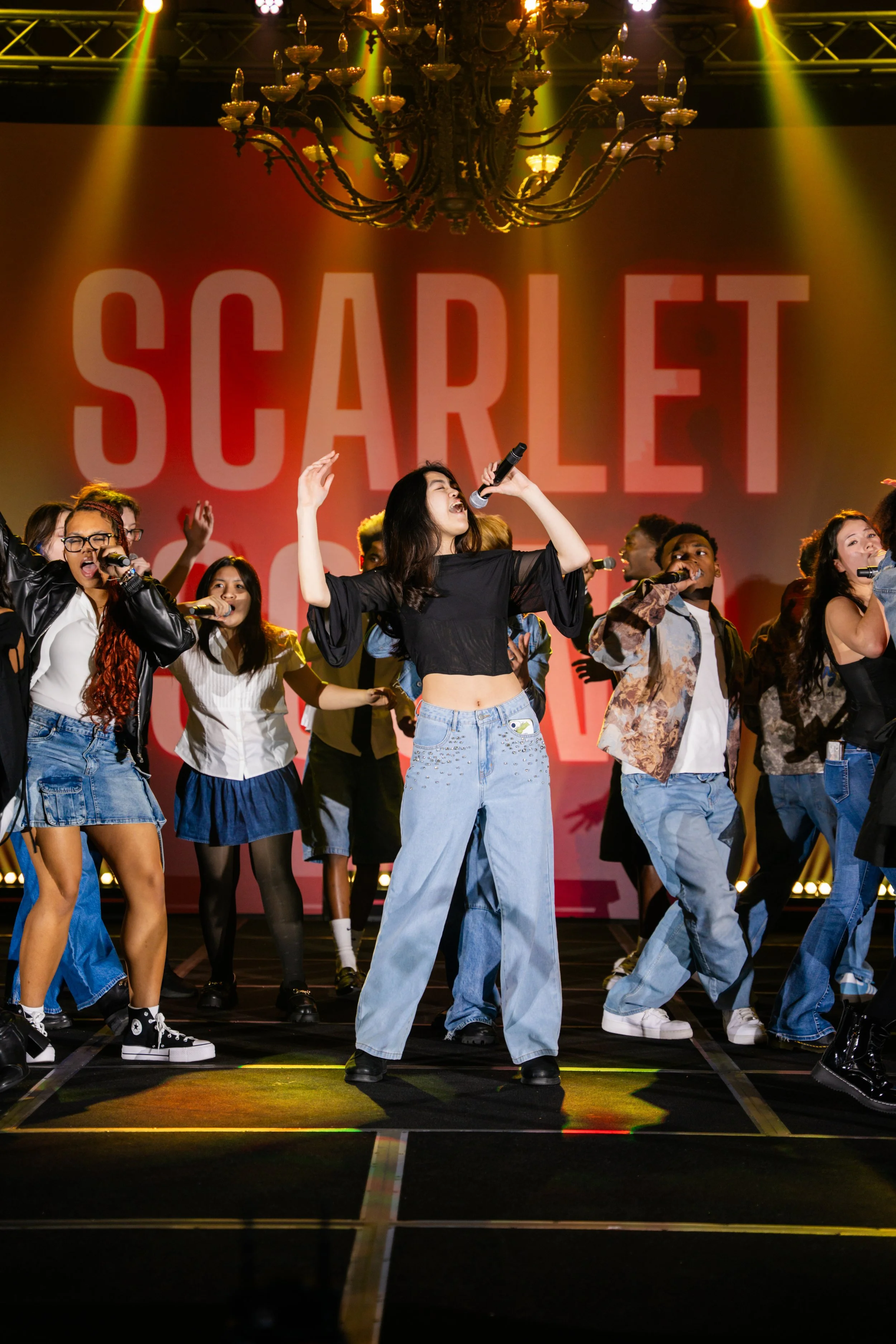 A group of diverse young people singing and dancing on stage during a performance, with a woman in the center holding a microphone and raising her arms, under colorful stage lighting and a large red screen in the background with the words "SCARLET".