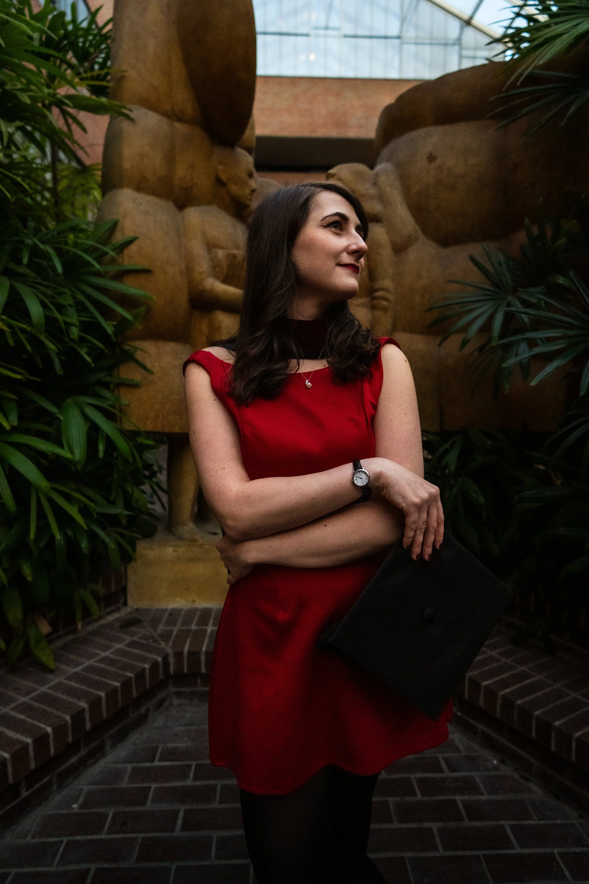 A woman in a red dress holding a black folder, standing in an indoor garden with large rocks and lush green plants.
