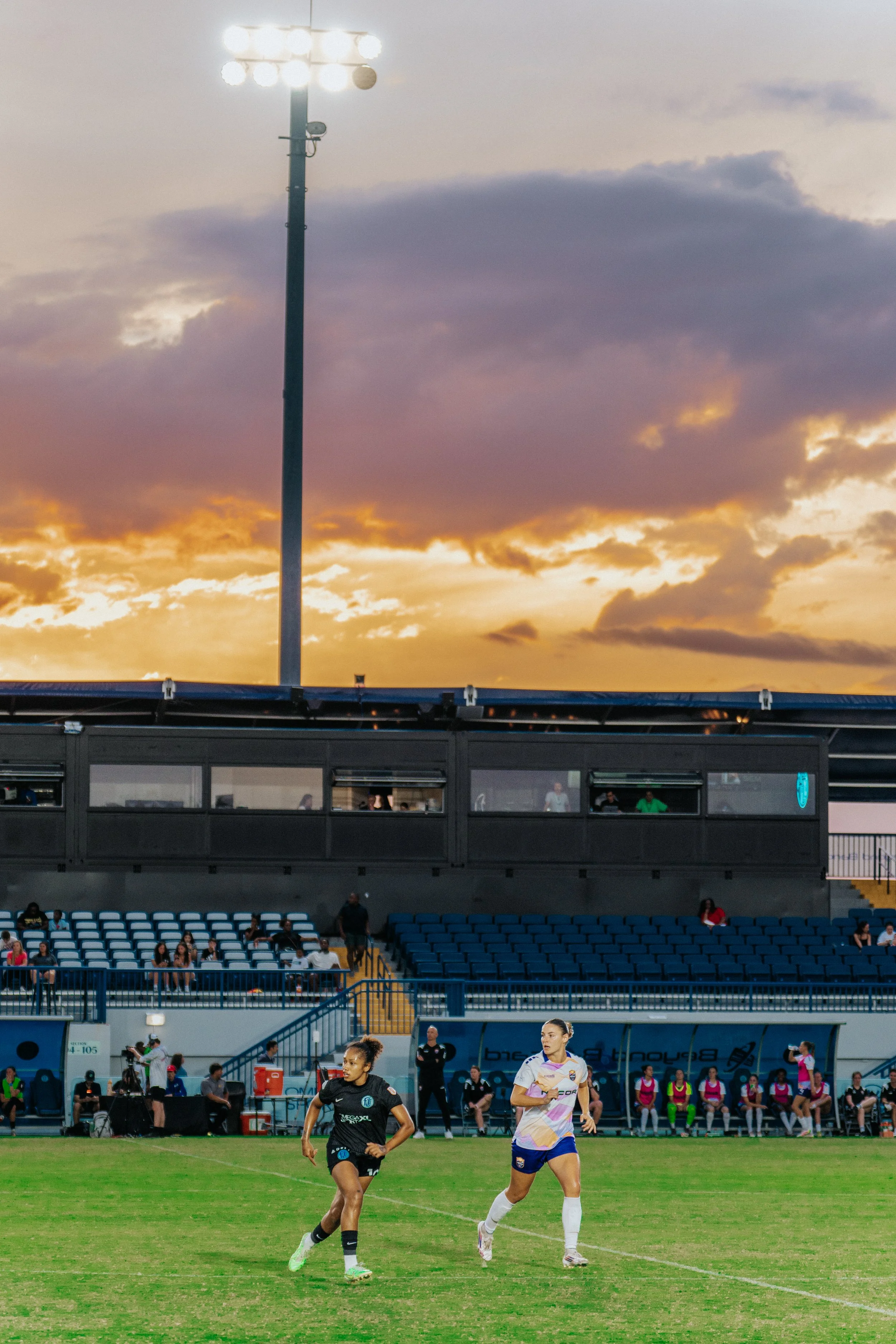 Women playing soccer on a field with spectators and a large stadium structure in the background during a sunset.