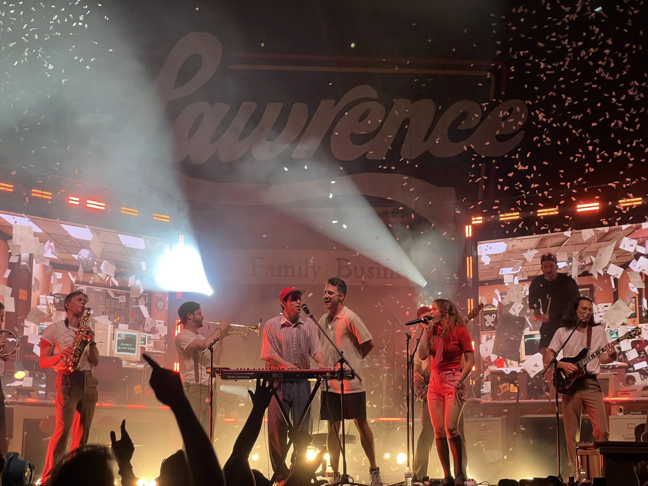 People performing live music on stage with a band, lights, and confetti, in front of a large sign that says 'Louvreence'.