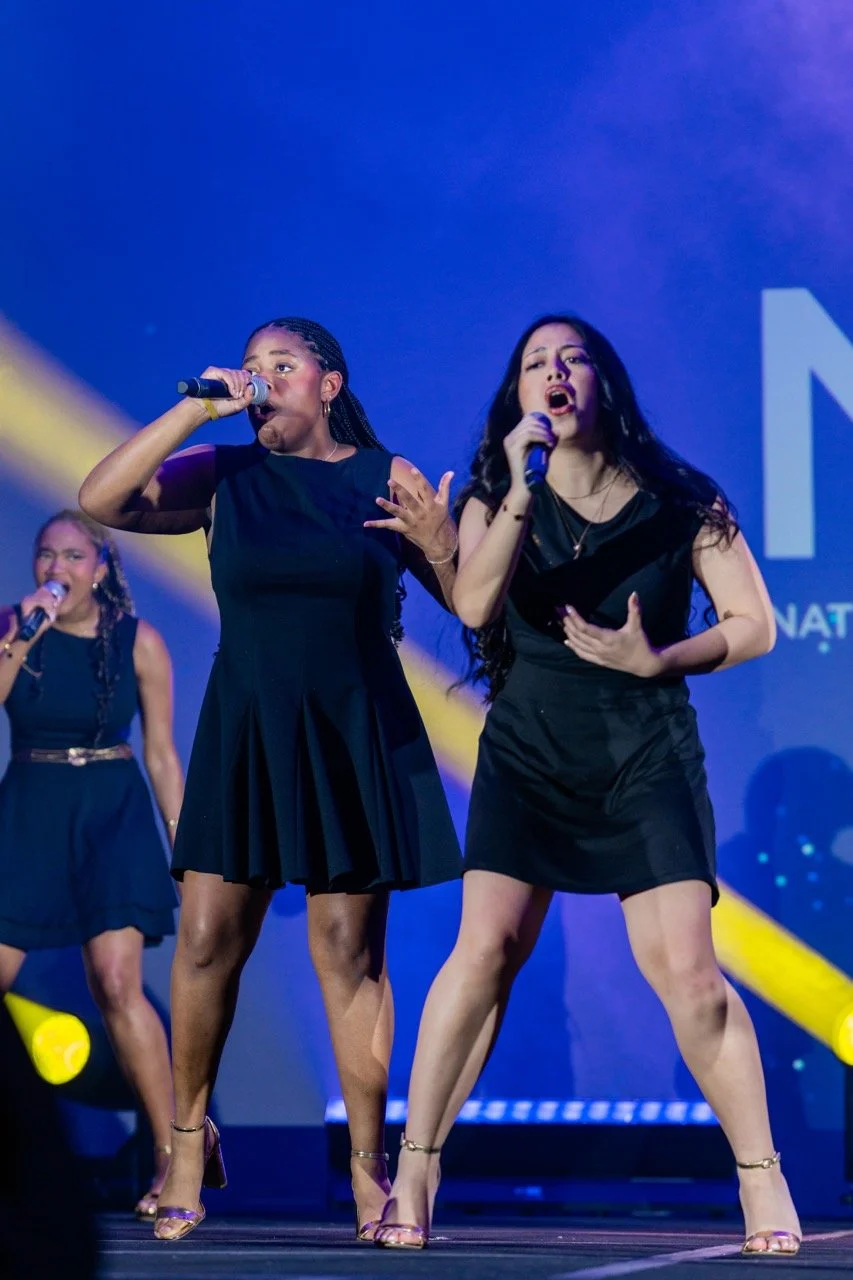 Three women singing on stage, holding microphones, with blue and yellow stage lighting.