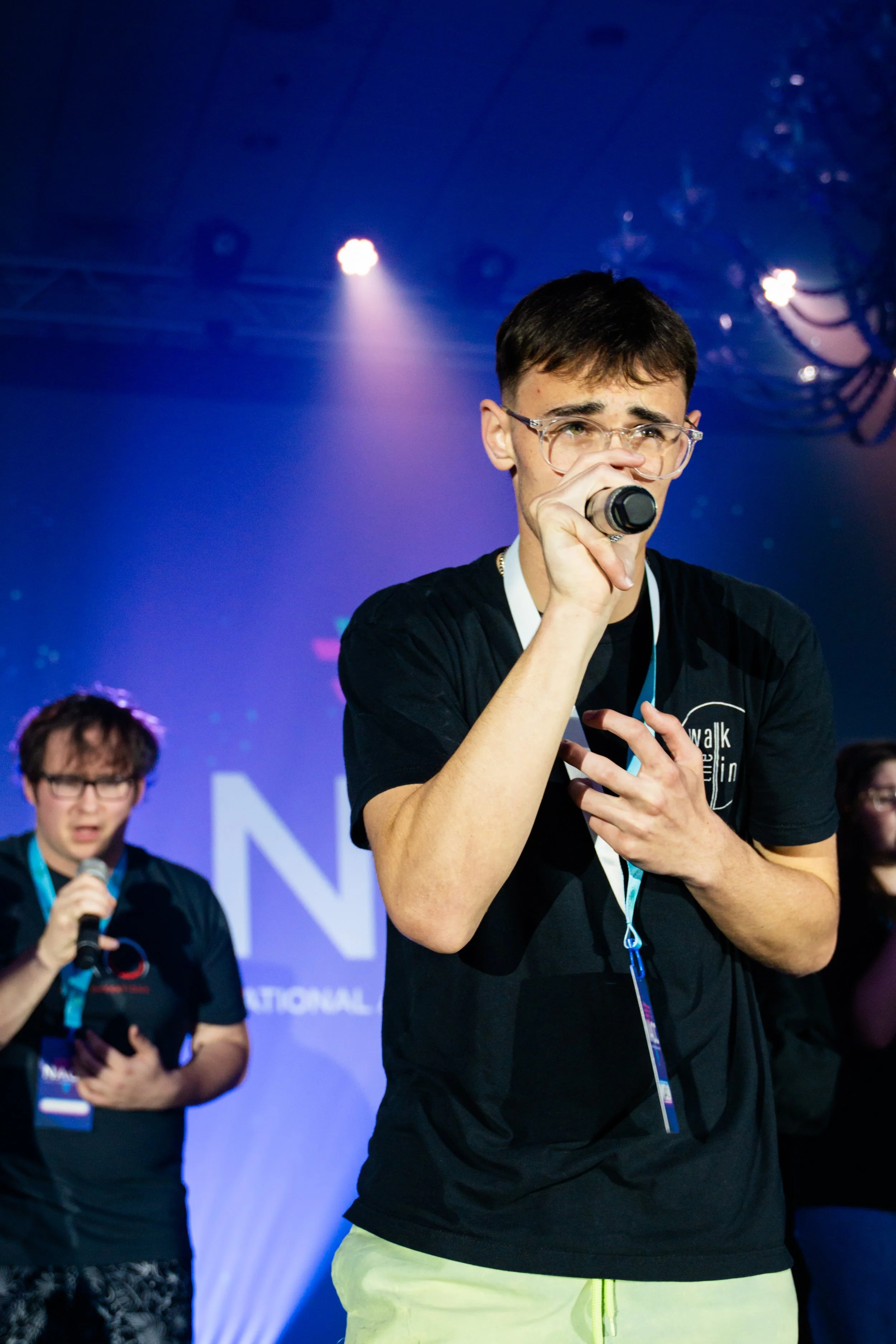 A young man with dark hair, glasses, and a black T-shirt holding a microphone on a stage, with another person in the background speaking into a microphone. Both appear to be participating in a public event or presentation, with a blue-lit backdrop.