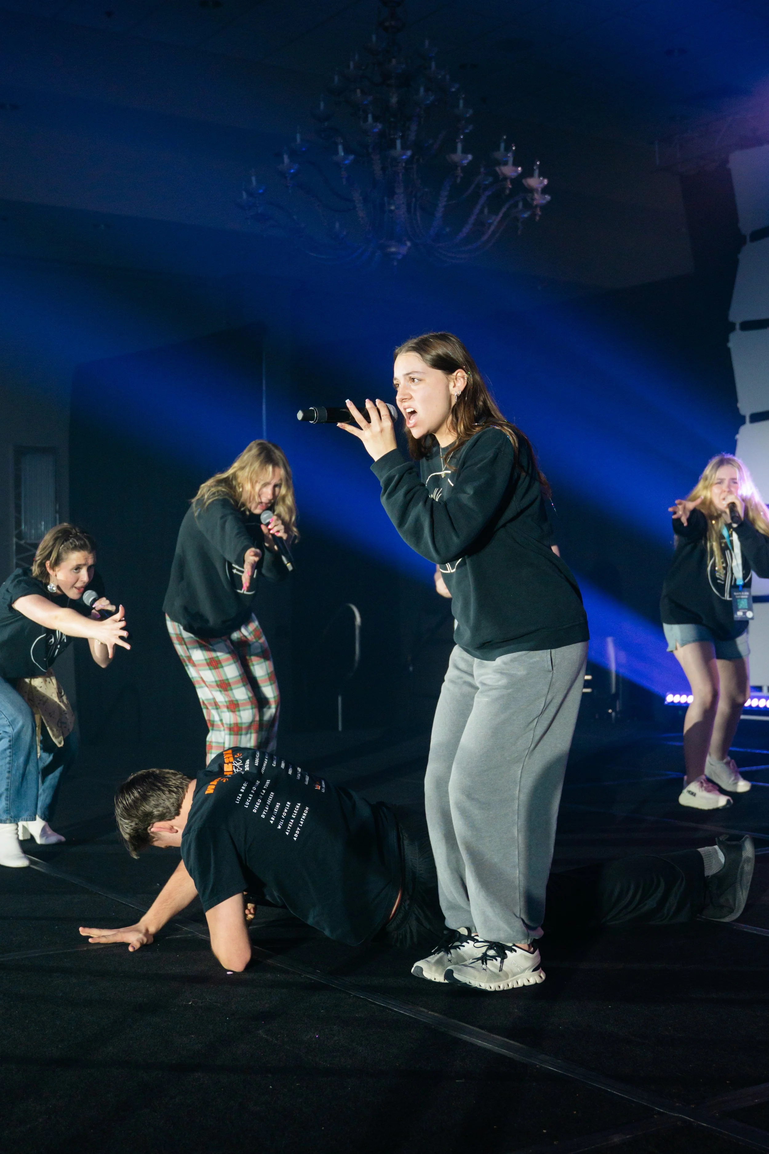 Young woman singing into a microphone on stage with backup dancers performing synchronized dance moves around her, stage lighting, and a chandelier visible in the background.