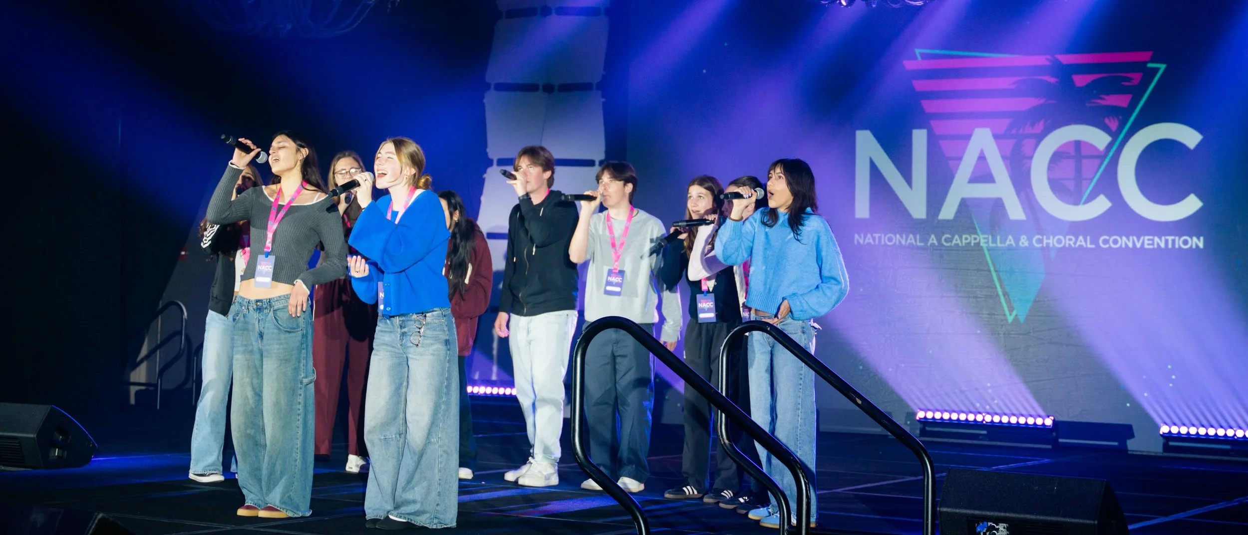 Group of young women singing on stage at the NACC National A Cappella & Choral Convention, with a large screen displaying the NACC logo in the background.