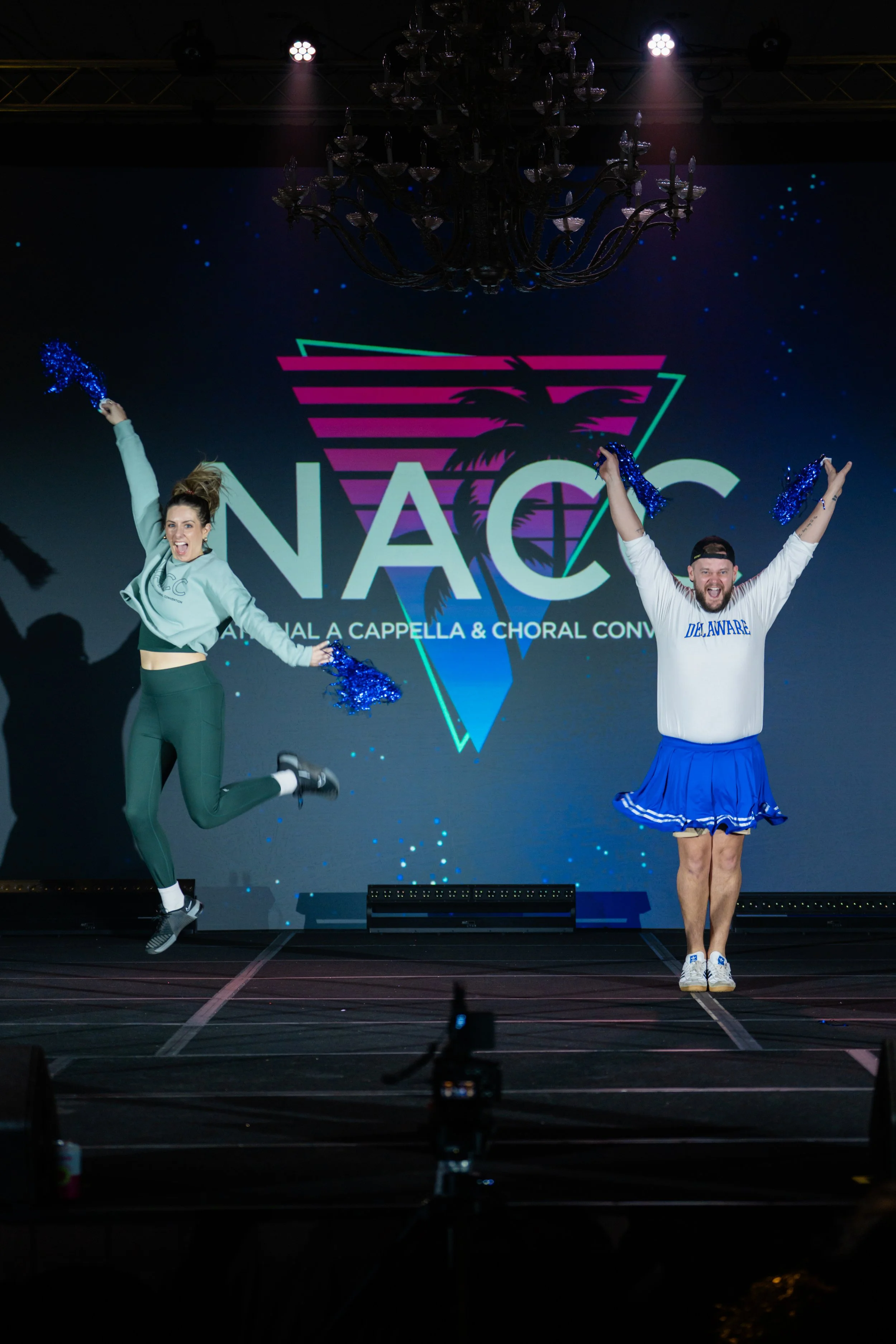 Two cheerleaders jumping on stage in front of a large screen displaying the NAC logo with a palm tree design. The woman on the left has her hair in a bun and is wearing a gray jacket and green pants. The man on the right is wearing a white sweatshirt