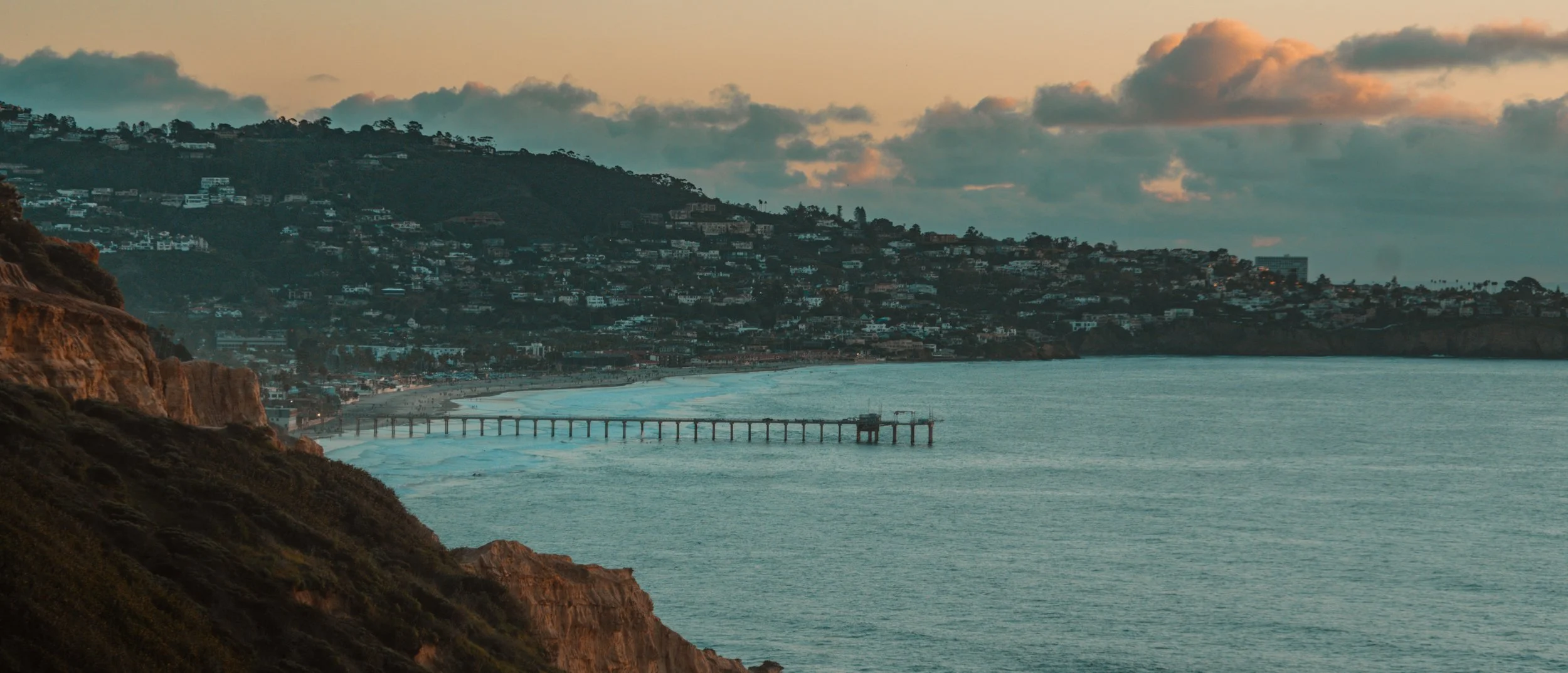 Scenic view of a beach with a pier extending into the ocean, rocky cliffs on the left, and a hillside with houses in the background at sunset.
