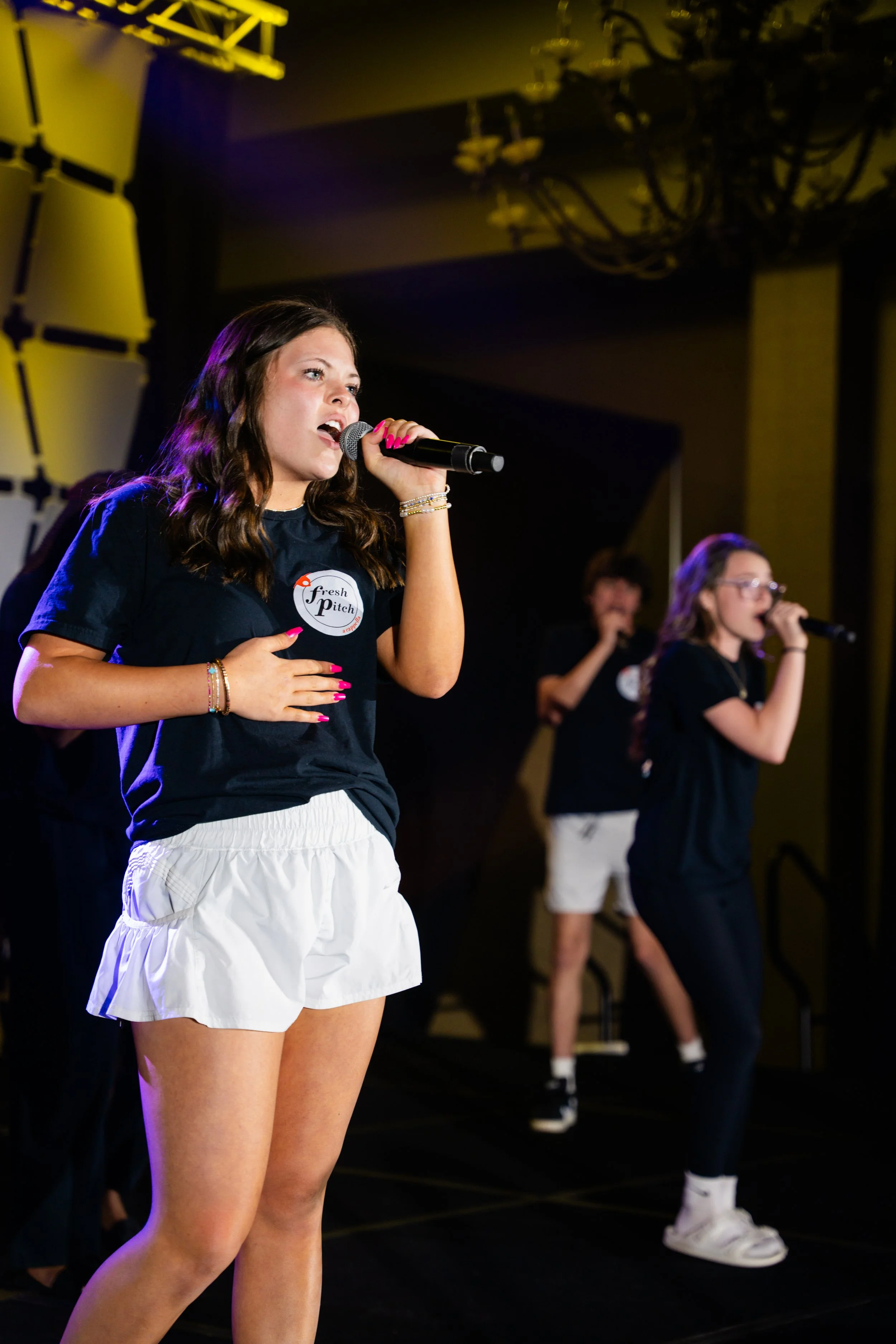 A young woman singing into a microphone during a performance, wearing a black T-shirt with the words 'fresh pitch' on it and white shorts. Other performers are visible in the background.