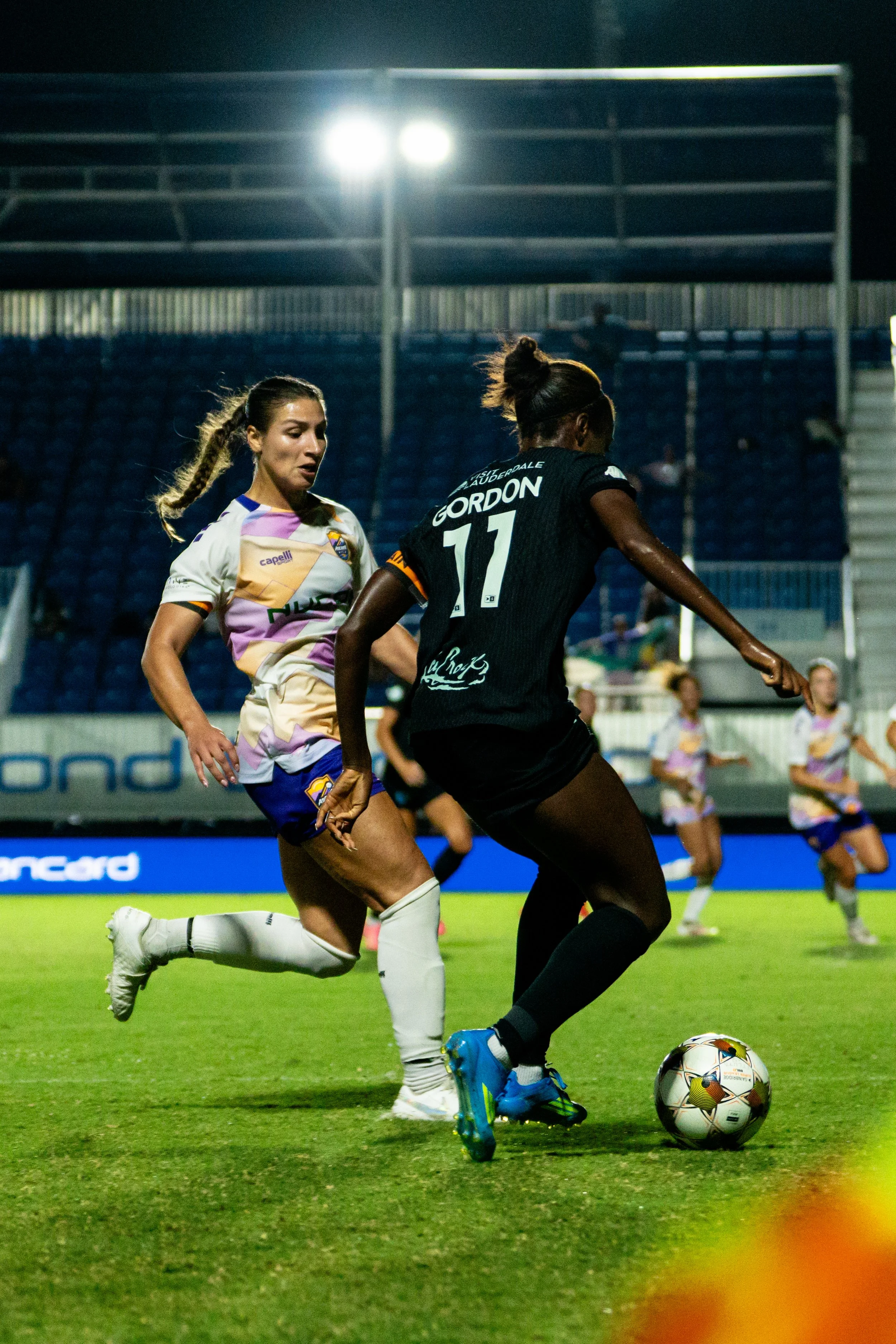 Two female soccer players competing for the ball on a green field at night under stadium lights, with one in a black uniform and the other in a multicolored uniform.