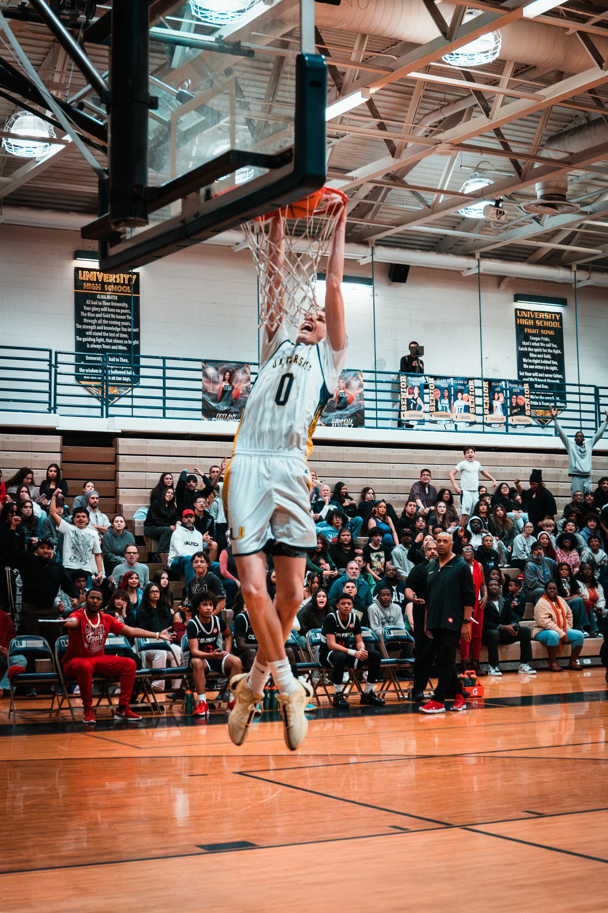A basketball player wearing a white jersey with the number 0 is dunking the ball into the hoop during a game in a gymnasium. There are spectators and team members seated and standing along the sidelines, and banners on the wall behind them.