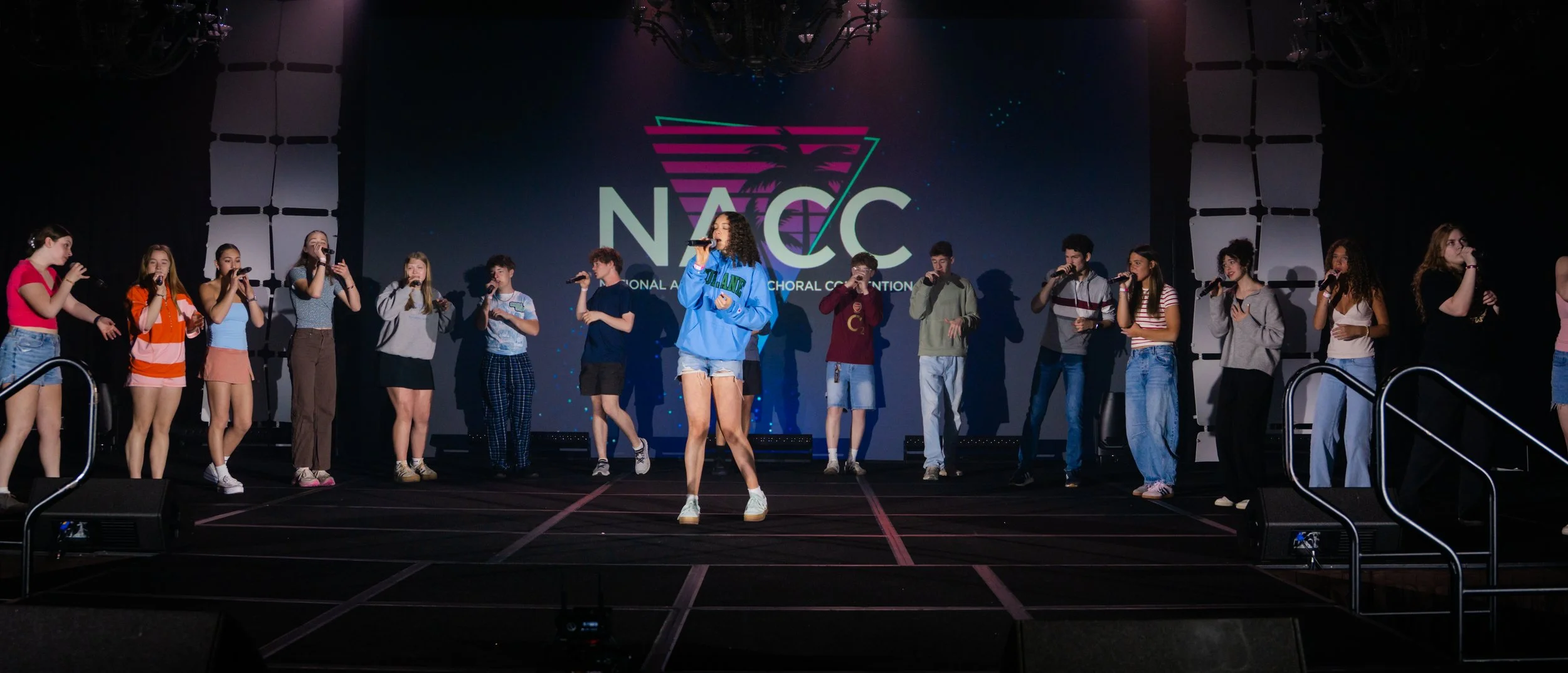 Group of teenagers performing on stage at the National Association of Choirs Conference, with a large screen showing the NAC logo and a palm tree in the background.