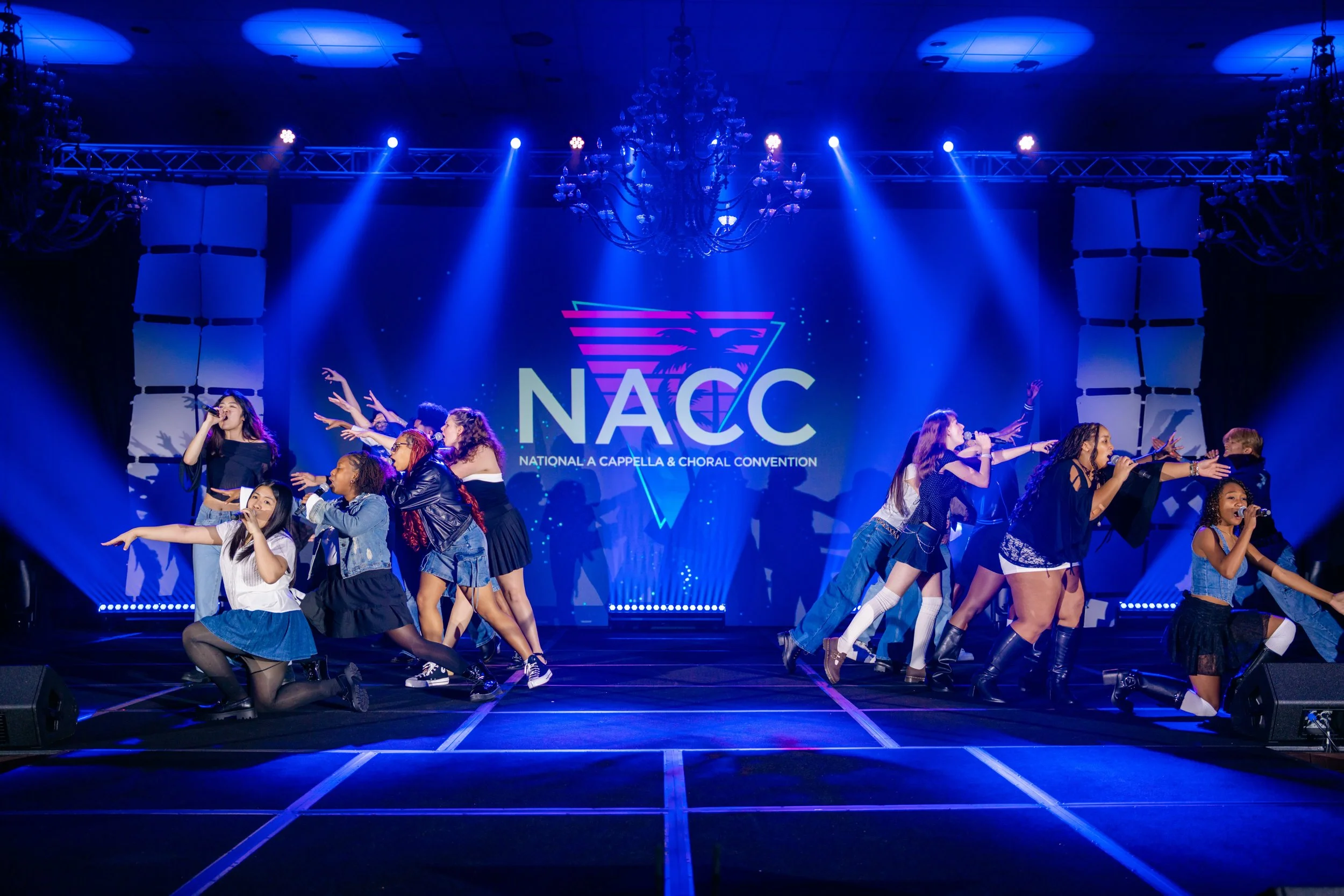 Group of diverse young women performing on stage at the National A Cappella & Choral Convention, with blue stage lighting and a large screen displaying the event's logo.