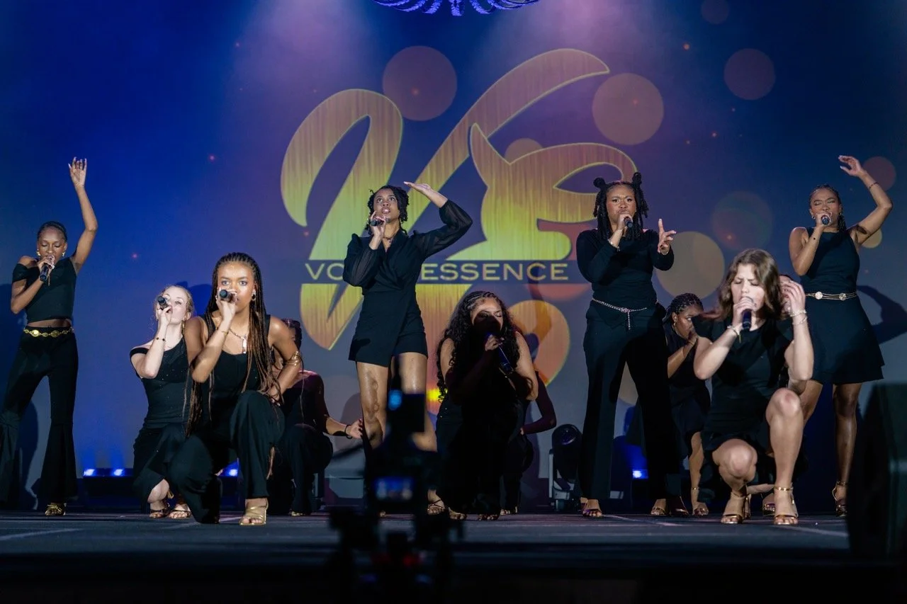 Group of women performing on stage at a VoxXessence event, six women standing and kneeling while singing into microphones, with a large VoxXessence logo in the background.