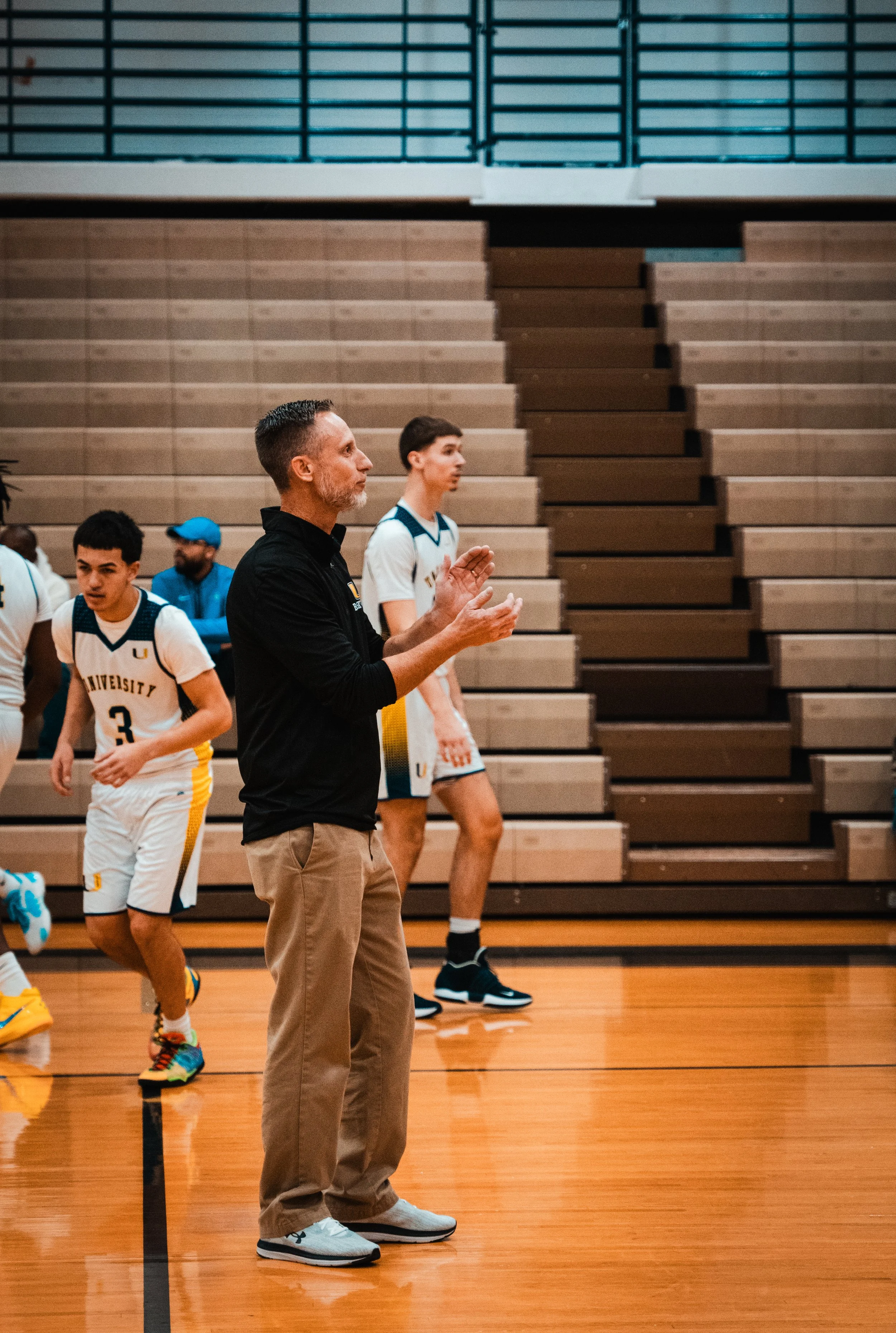 Basketball coach clapping on the sideline of an indoor game, with players in white and yellow uniforms in the background.