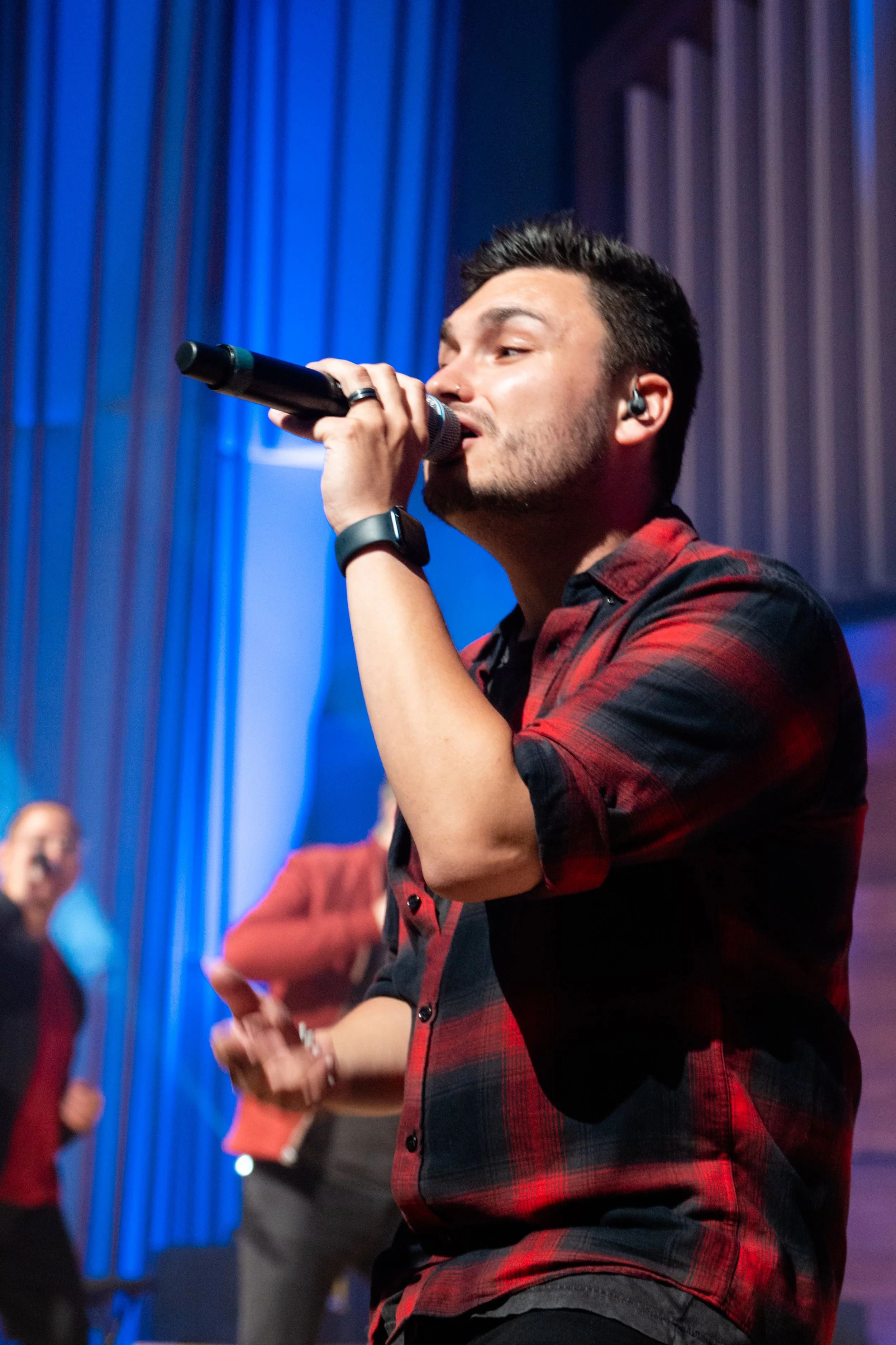 A young man with dark hair and a beard is singing into a microphone during a live performance. He is wearing a red and black plaid shirt and a smartwatch on his left wrist. The background features a stage with blue lighting and curtains, and a woman 
