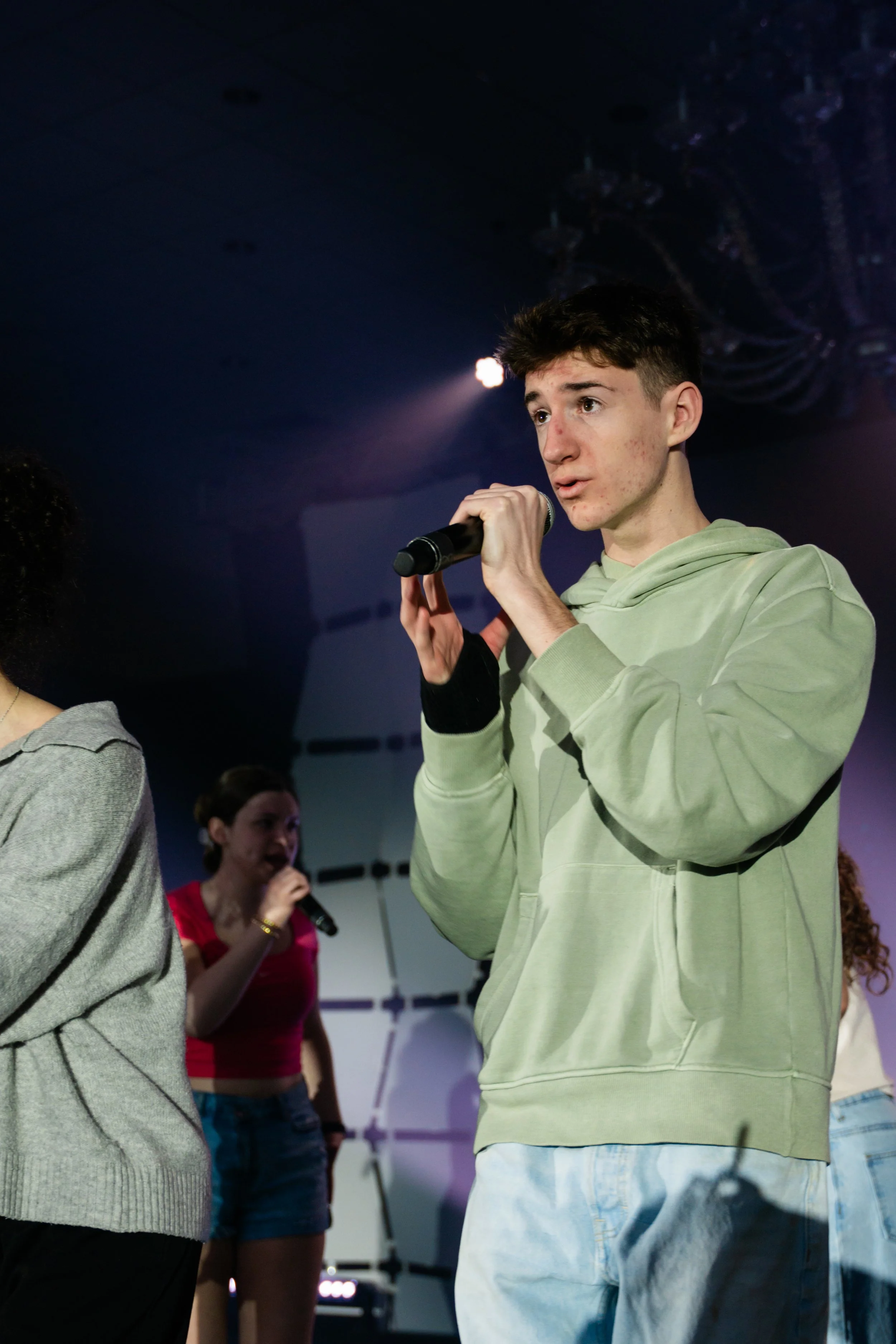 Young man holding a microphone, speaking or singing at an indoor event, with a dark background and other people, including a woman in a pink shirt holding a microphone, standing behind him.