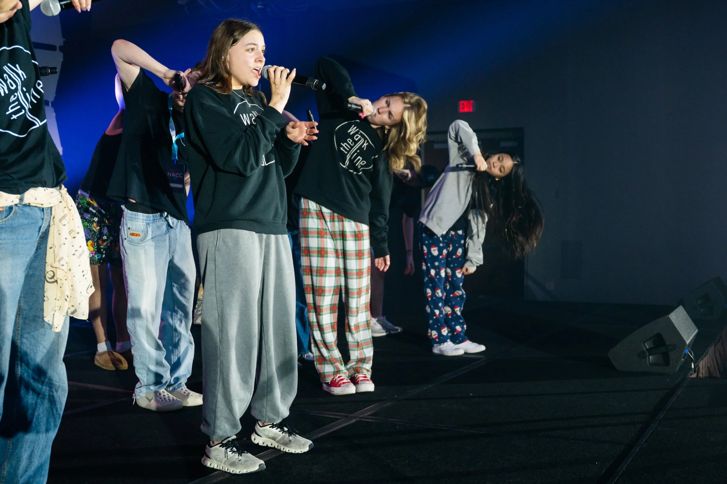 A group of young women standing on stage, some holding microphones, wearing casual and festive pajamas, possibly during a performance or event, with a dark background and stage lighting.