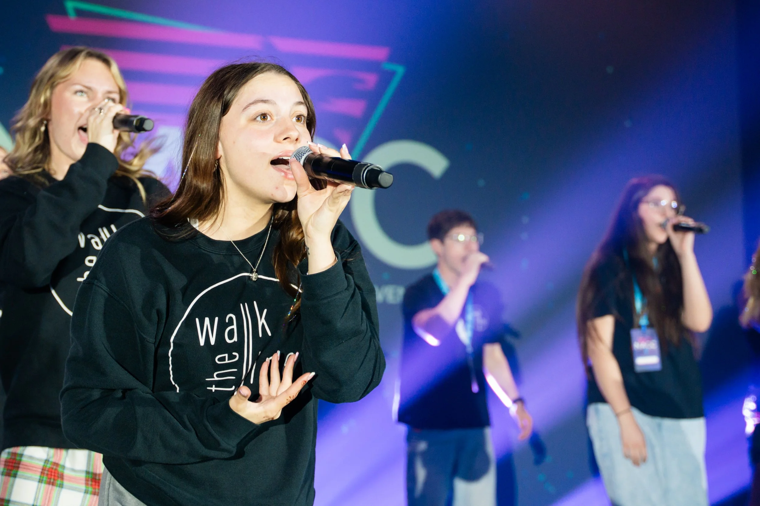 Group of young women singing into microphones on stage with colorful background lights.