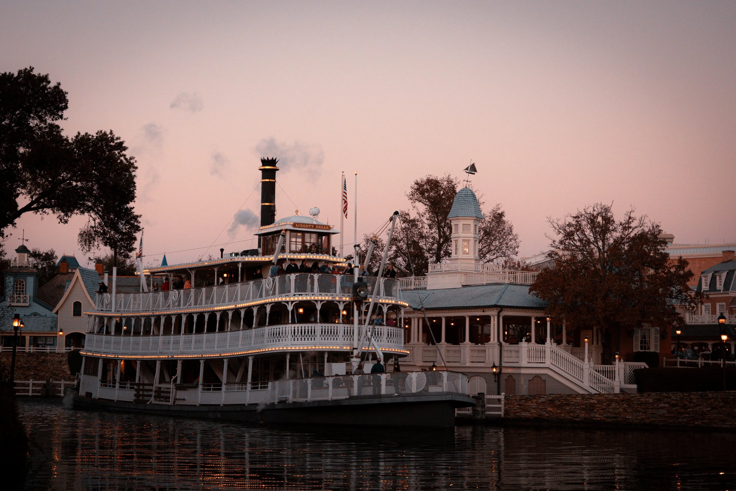 A large white paddlewheel boat docked by a river, with smoking chimney, lights, and a deck filled with people, set against a sunset sky with trees and historic buildings in the background.