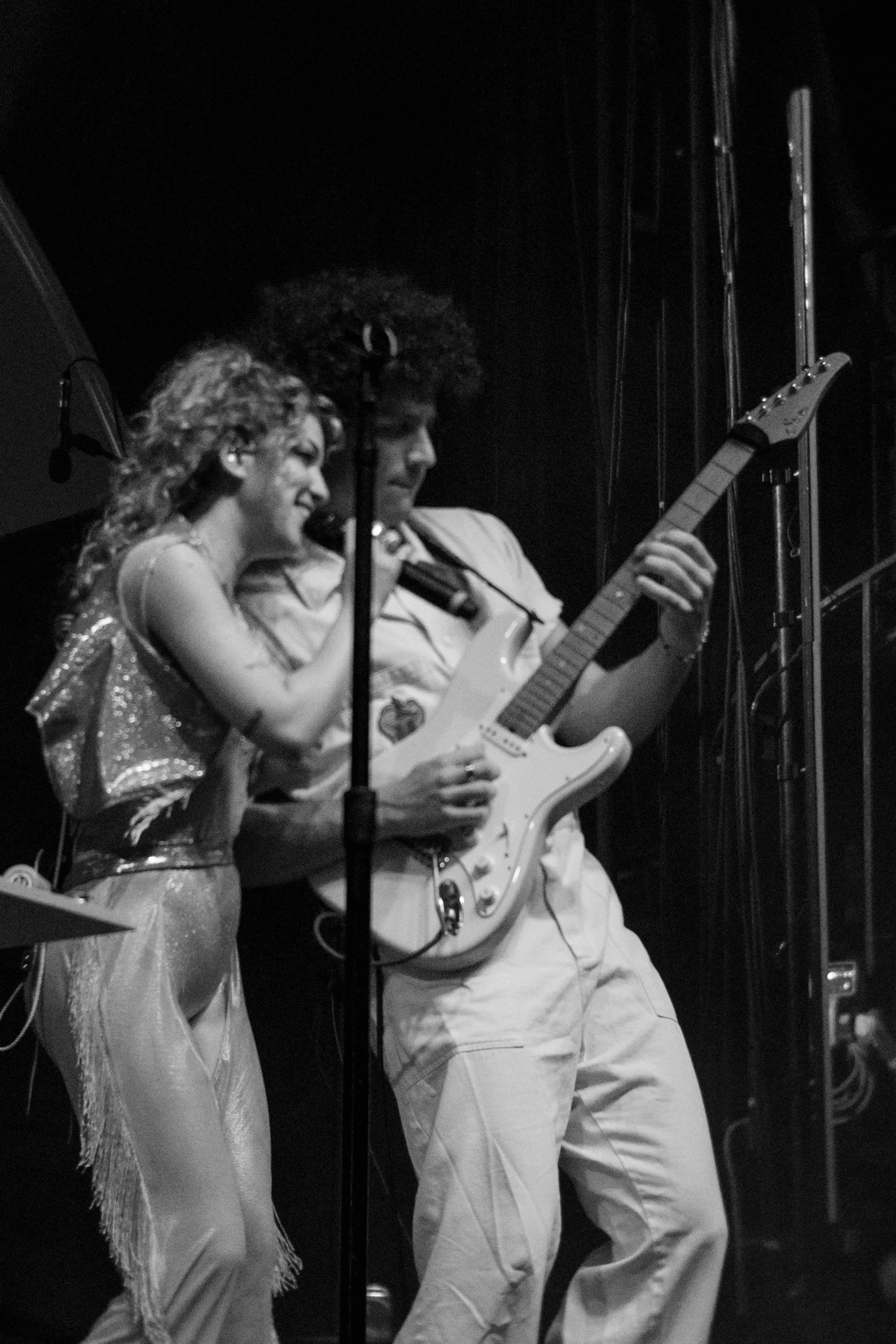 Two musicians, a woman singing into a microphone and a man playing an electric guitar, on stage during a performance. The image is in black and white.