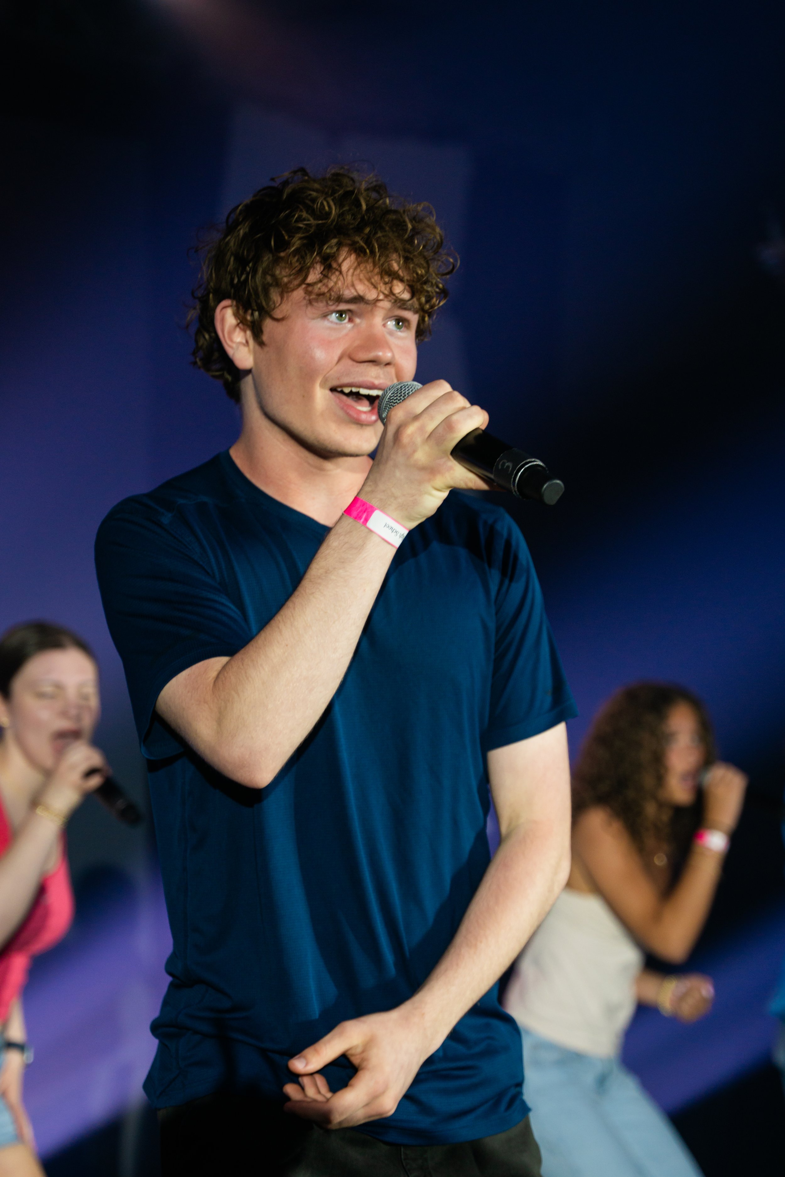 Young man with curly hair and a blue T-shirt holding a microphone, smiling on stage. In the background, women with microphones are singing or speaking, all appearing joyful and engaged.
