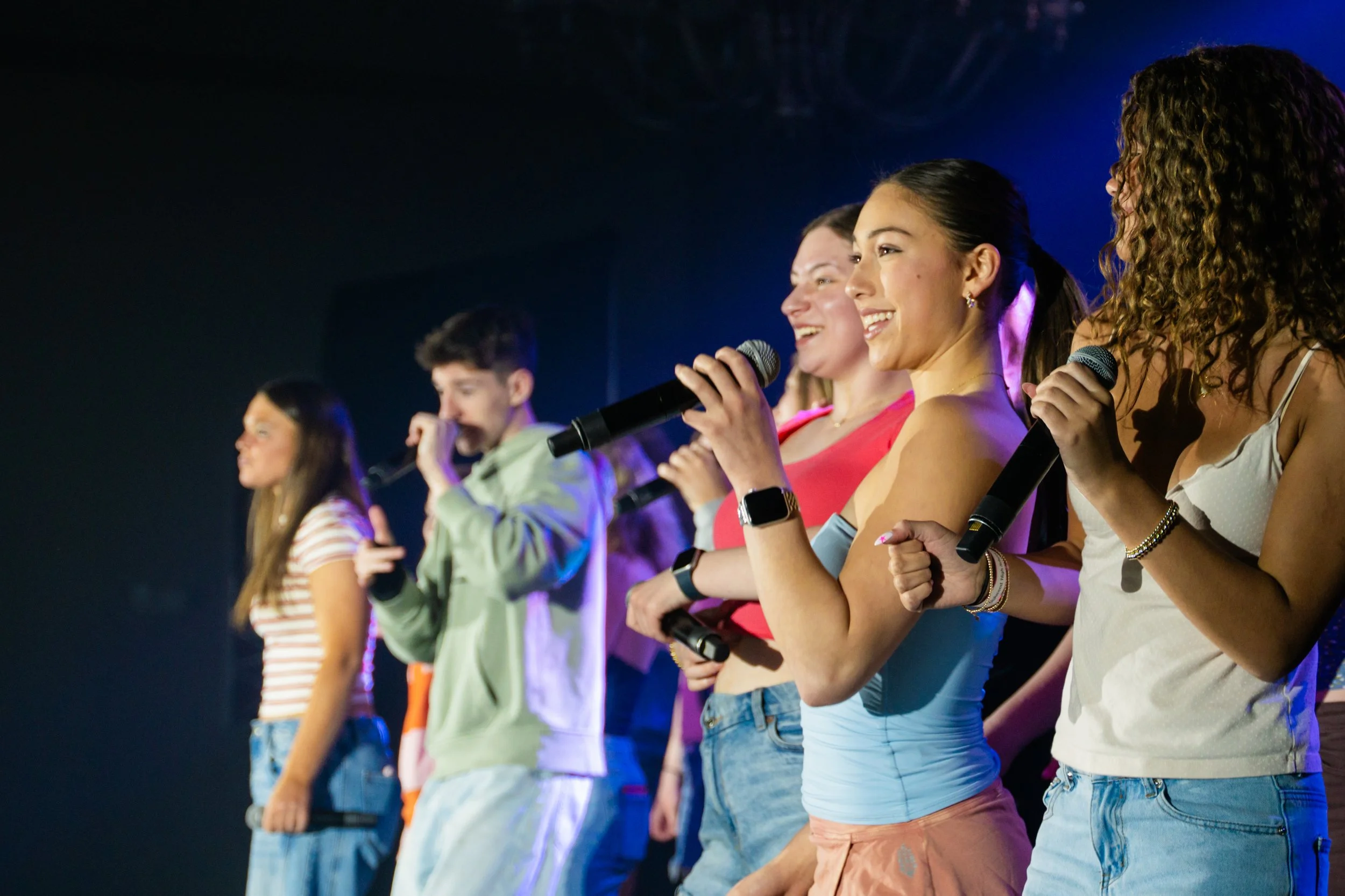 Group of diverse young people standing in a line, some holding microphones, on a stage during a performance or event, with dark background and stage lighting.