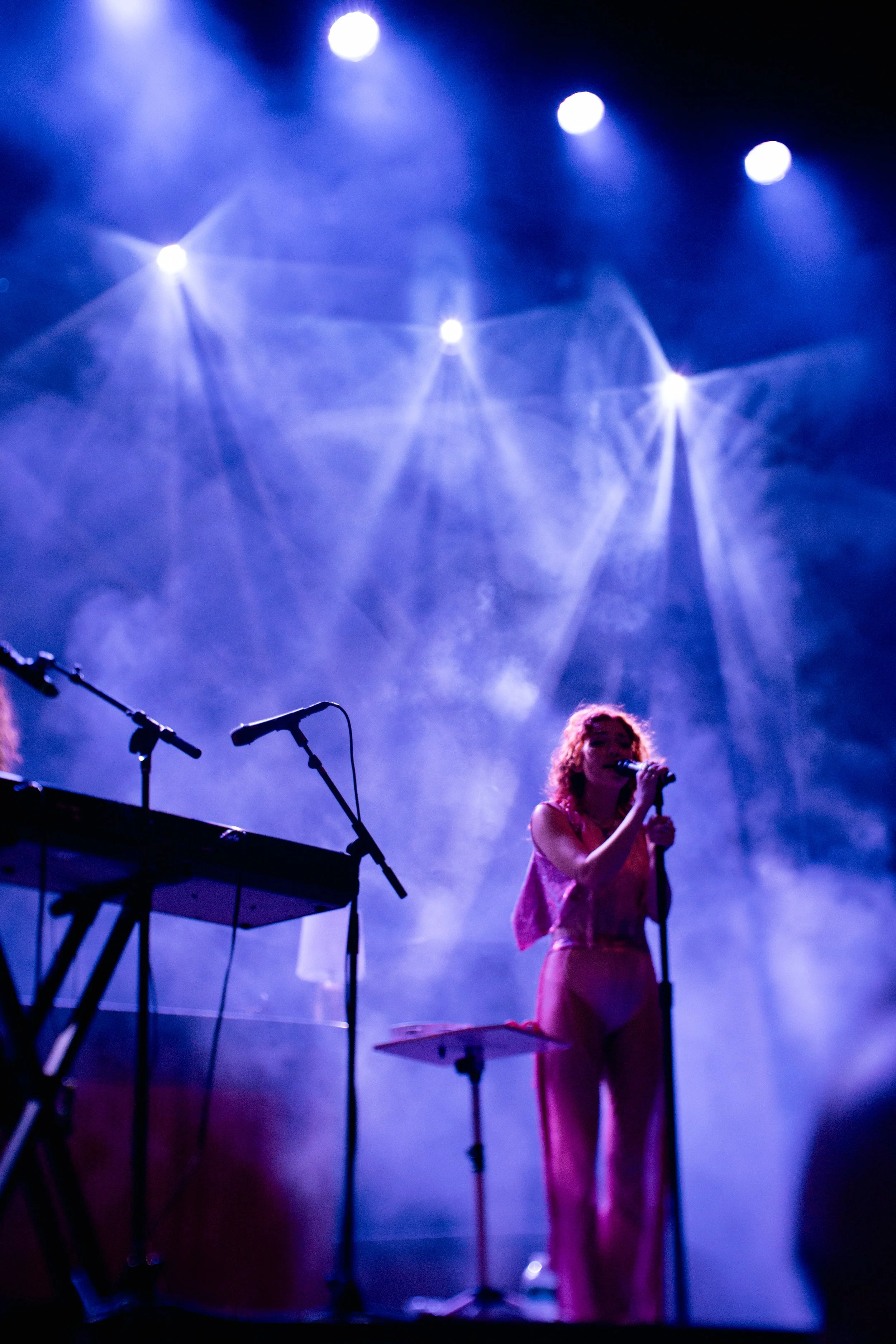 A singer with red hair on stage, wearing a pink outfit, holding a microphone, with a keyboard and microphone stand nearby, performing under blue stage lights.