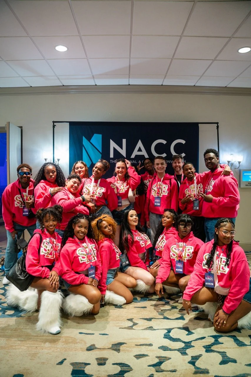 Group of young women and men posing for a photo at the NACAC event, wearing matching pink hoodies with SIE written on them, some with medals around their necks.
