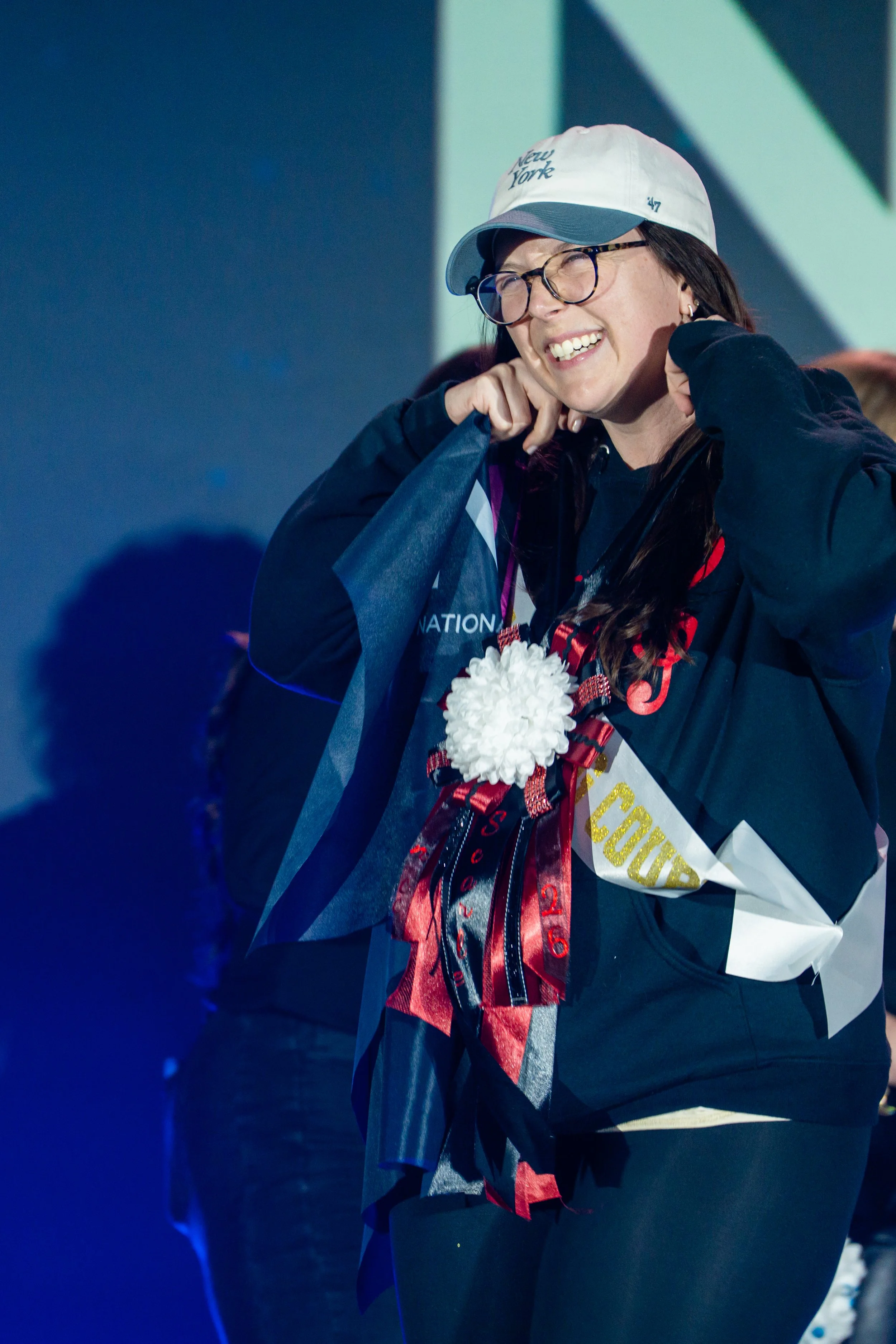 A smiling woman is wearing a white baseball cap with 'New York' written on it, glasses, and a dark jacket. She has a floral corsage and is holding a sash that says 'Winner 2023'. She appears to be at a celebration or pageant event.