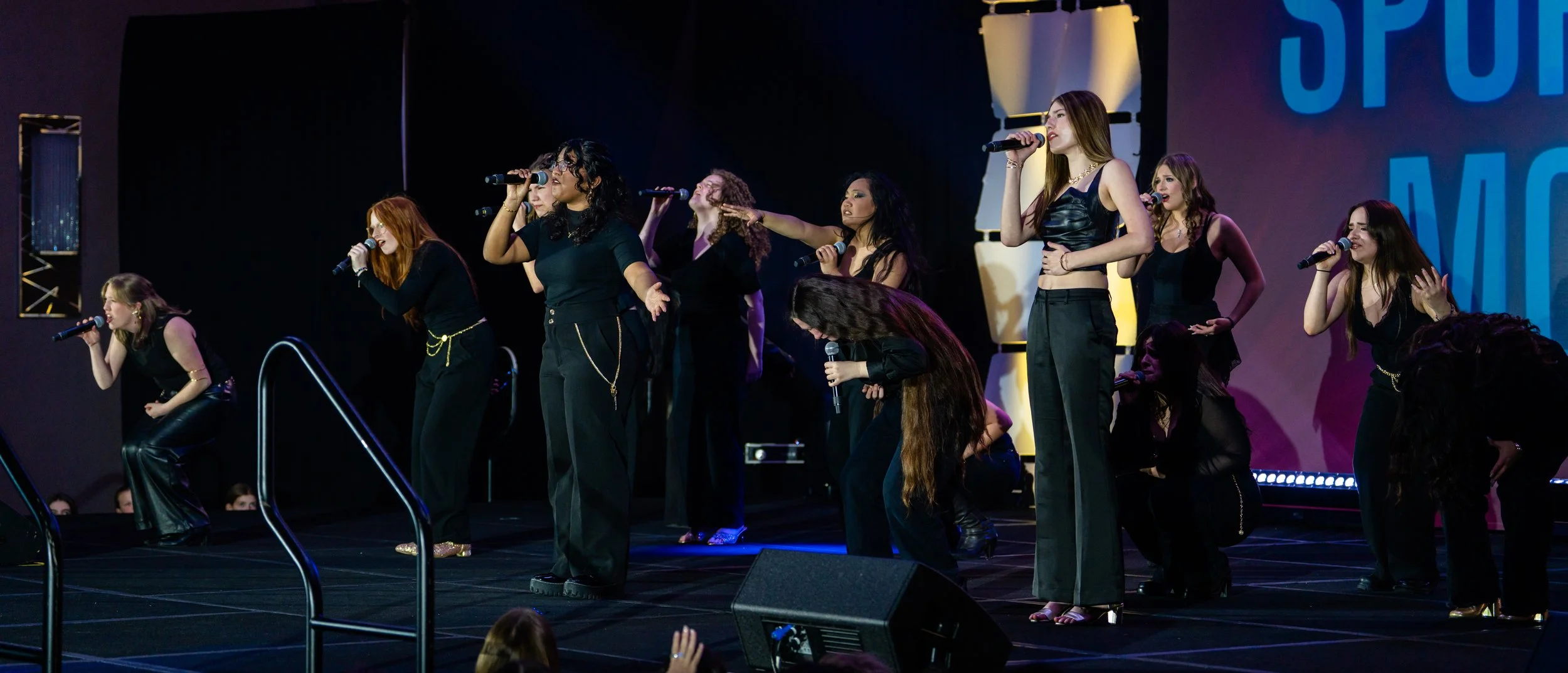 A group of female singers performing on stage with microphones, dressed in black outfits, some kneeling and some standing, with a large screen background.