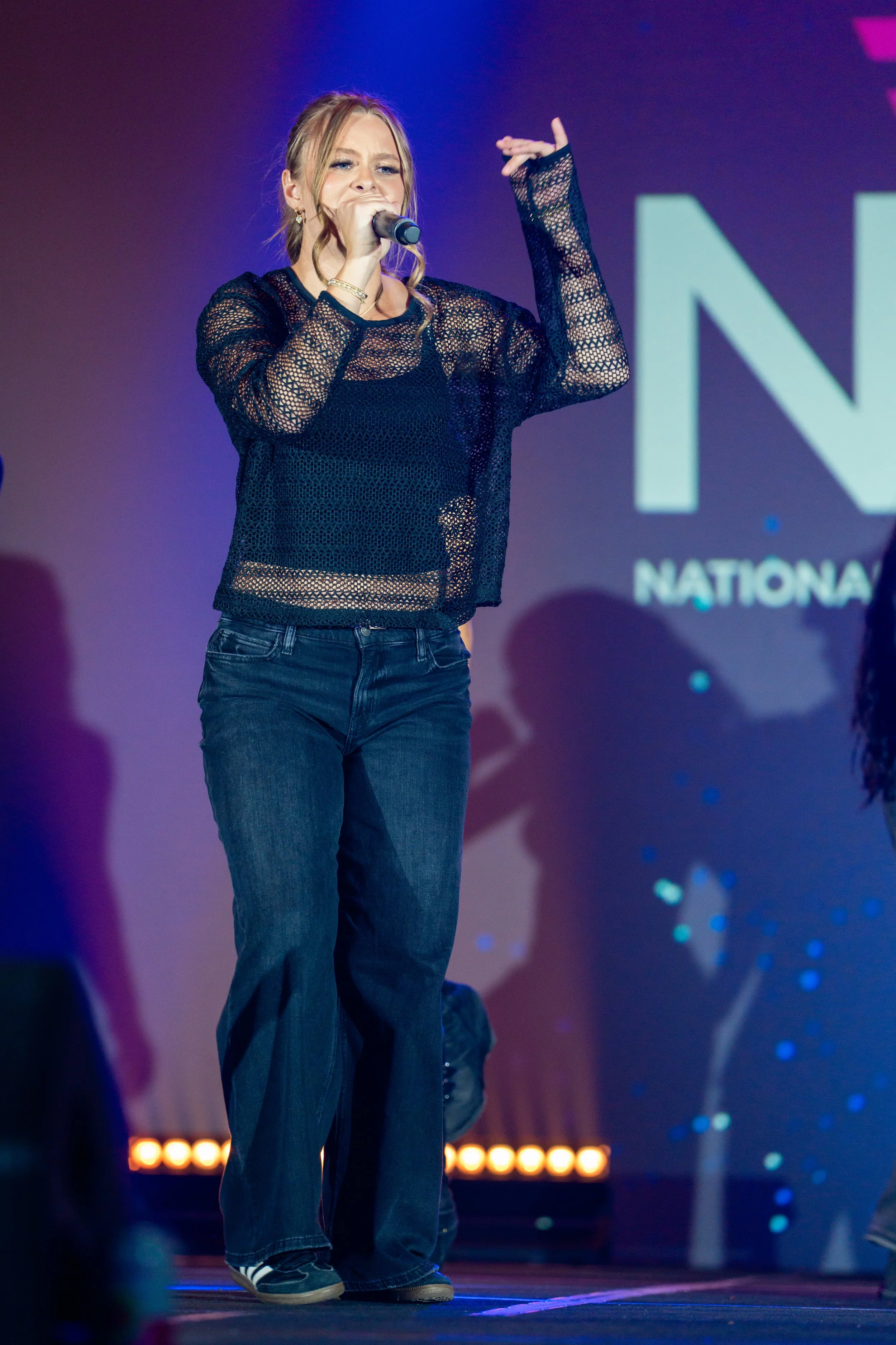 A young woman with blonde hair singing into a microphone on stage, wearing a black mesh long-sleeve shirt and blue jeans.