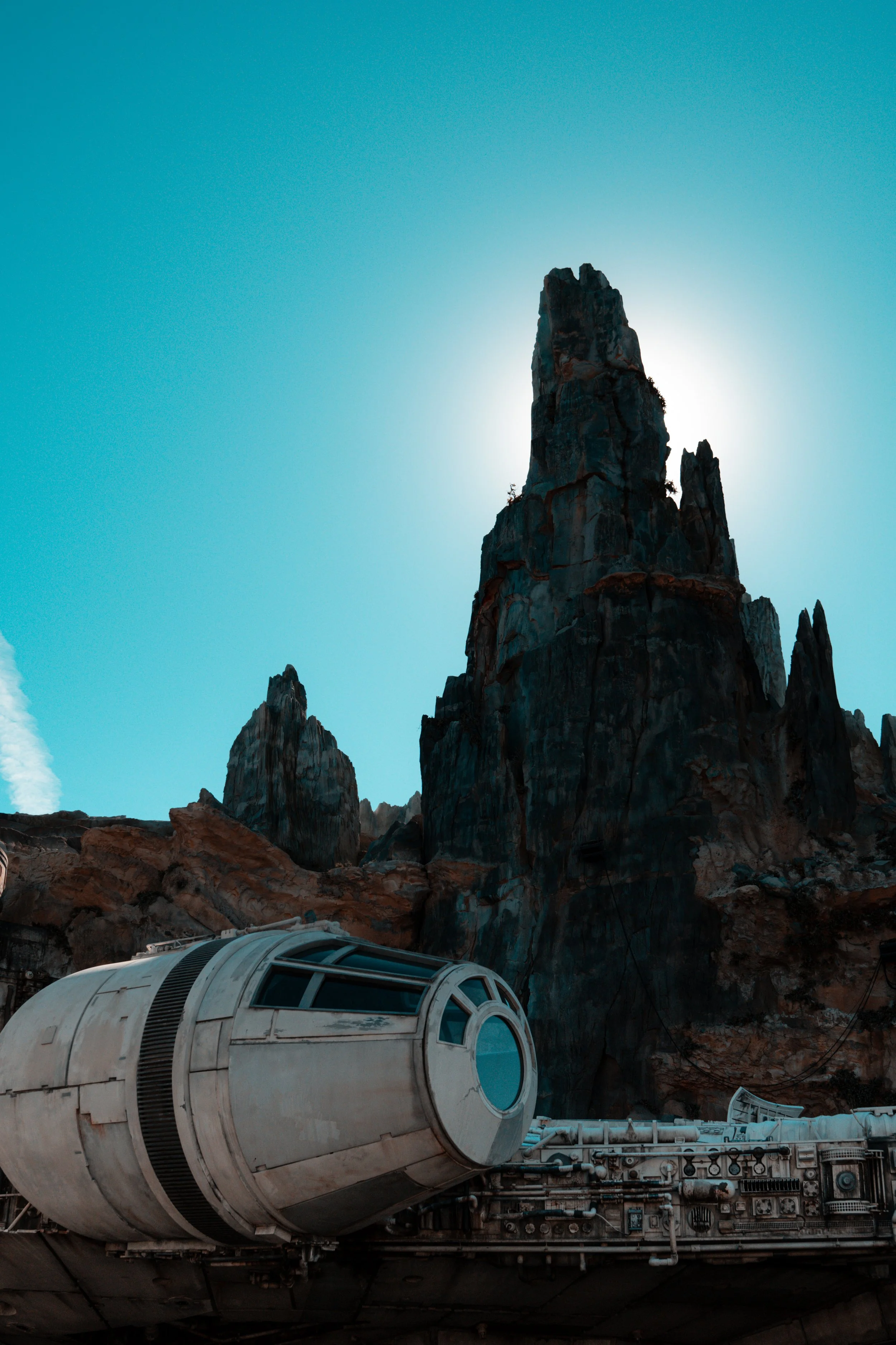 A scene from a science fiction setting featuring a spaceship docked at a base in a rocky, mountainous landscape with large, jagged rock formations and a bright sun behind one of the peaks.