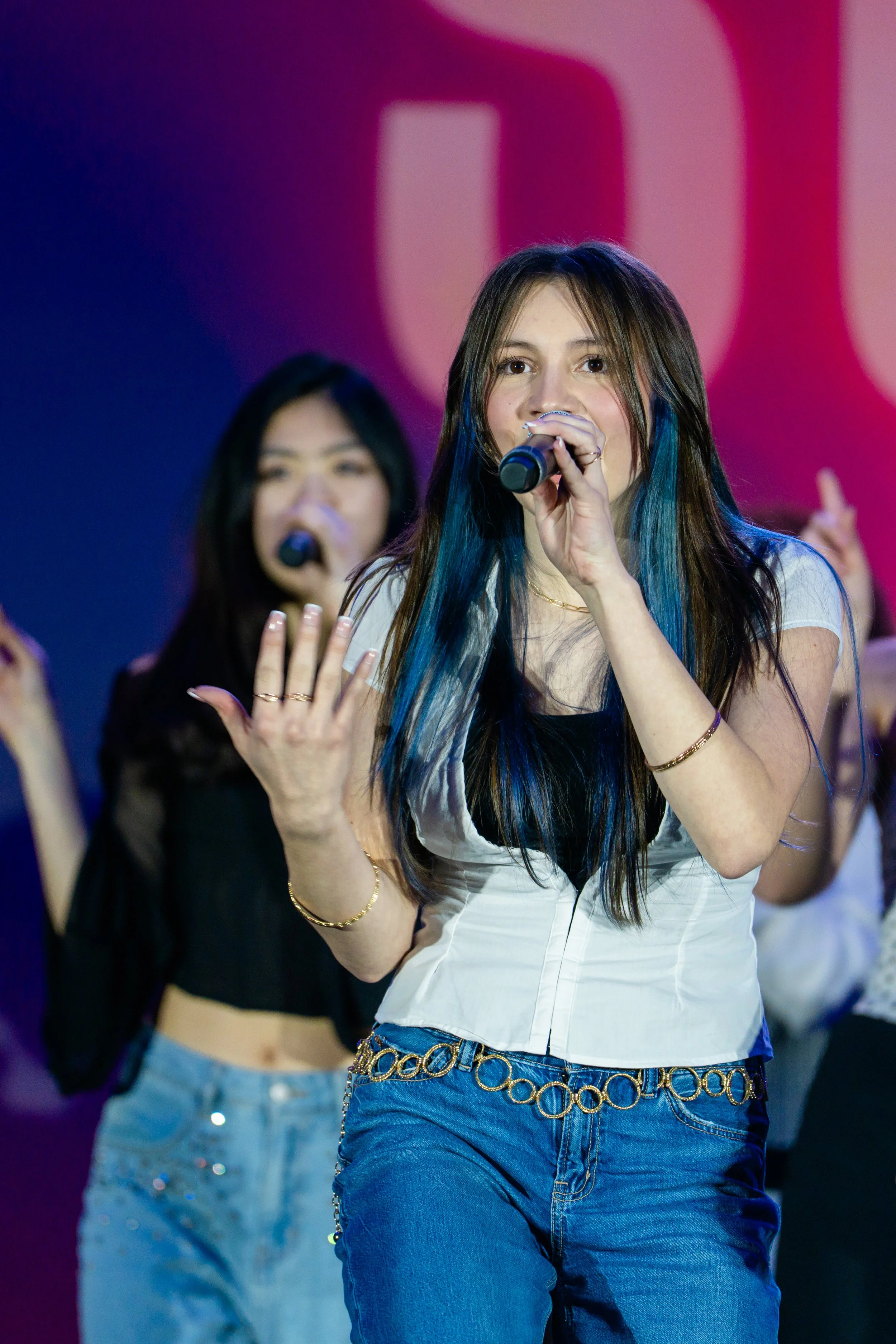 Teenage girl with long hair, wearing a white top and blue jeans, singing into a microphone on stage. A background dancer in black with a microphone is also visible.