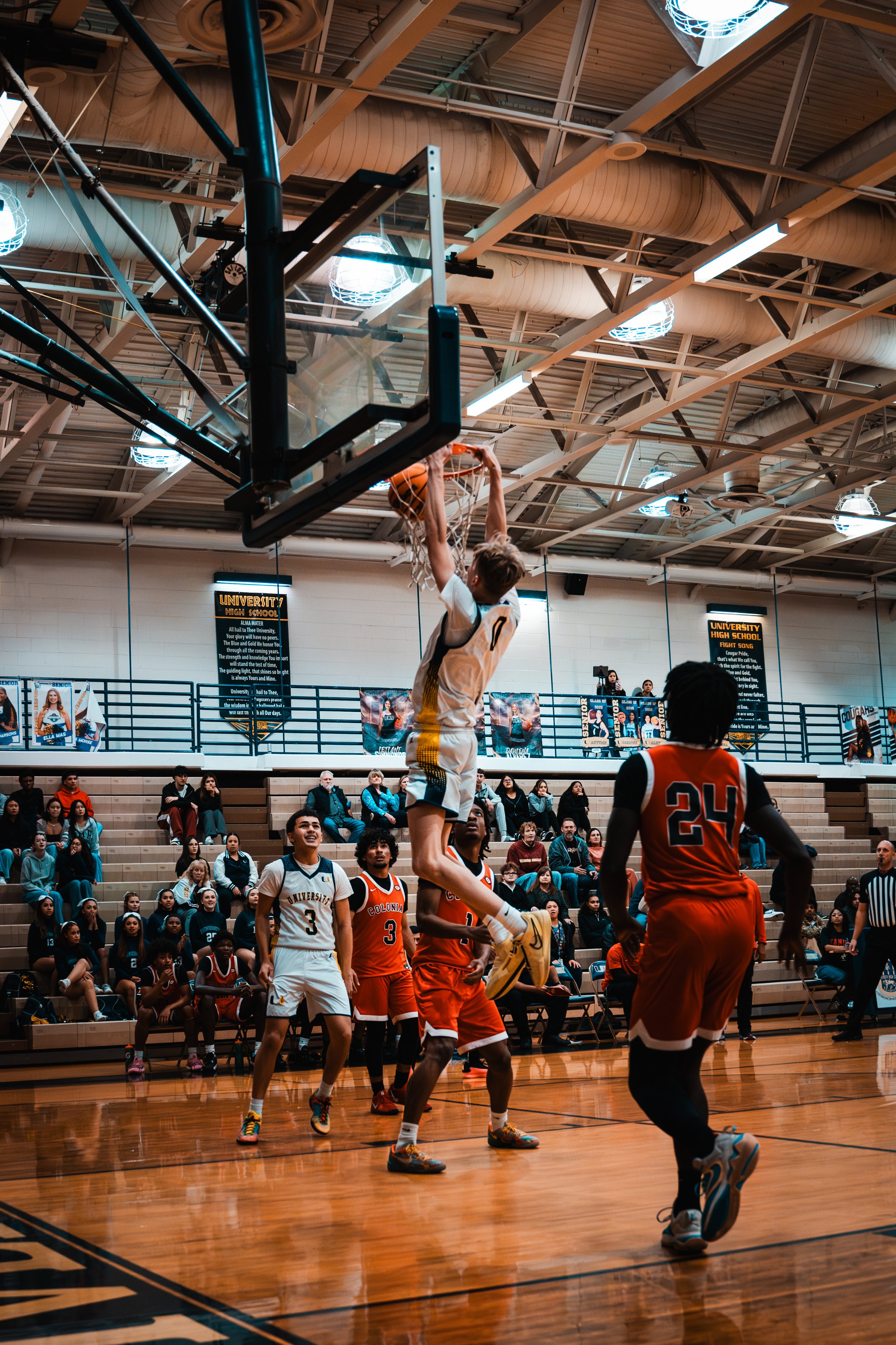 A basketball game in progress with a player jumping to score a basket while others watch. Spectators seated in the background.