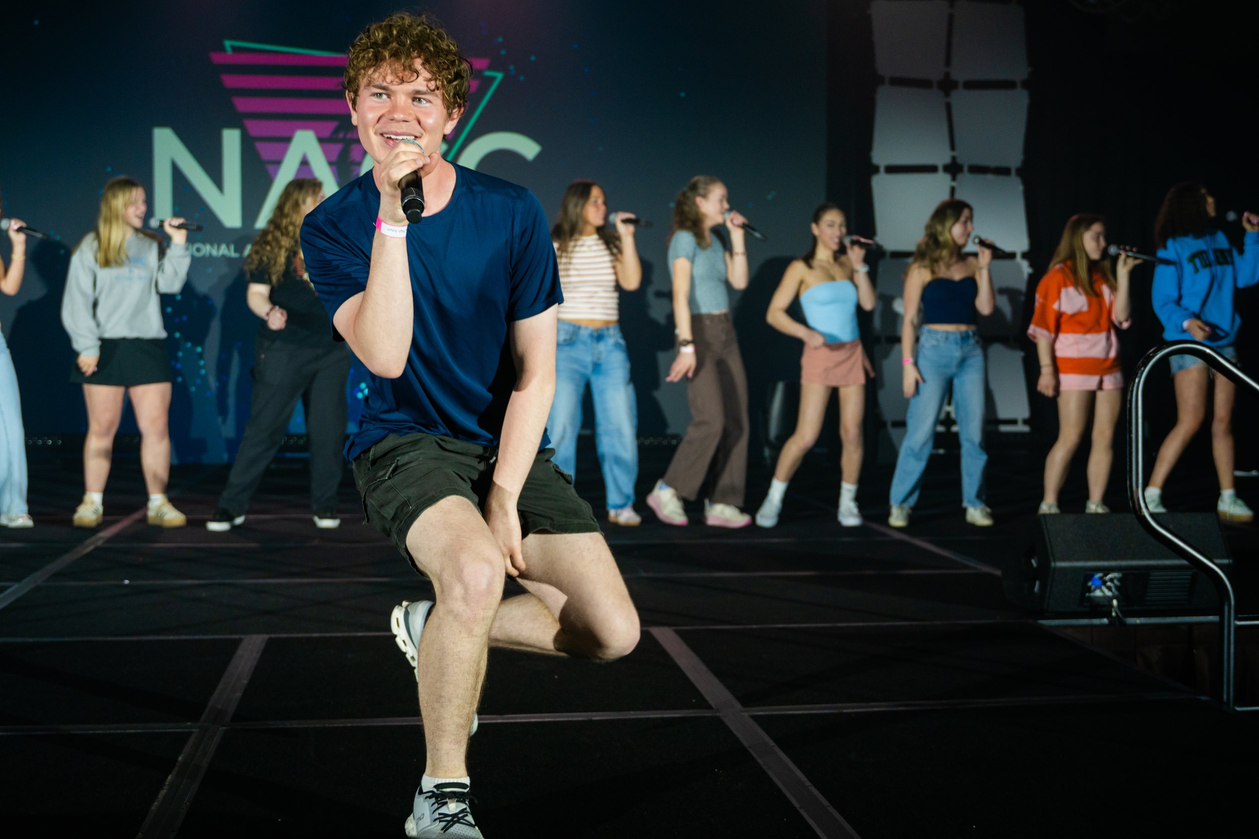 A young man with curly hair and a blue shirt is kneeling on stage, holding a microphone and smiling. Behind him, there is a group of girls standing in a line, each holding a microphone and singing. The background features a large illuminated logo tha