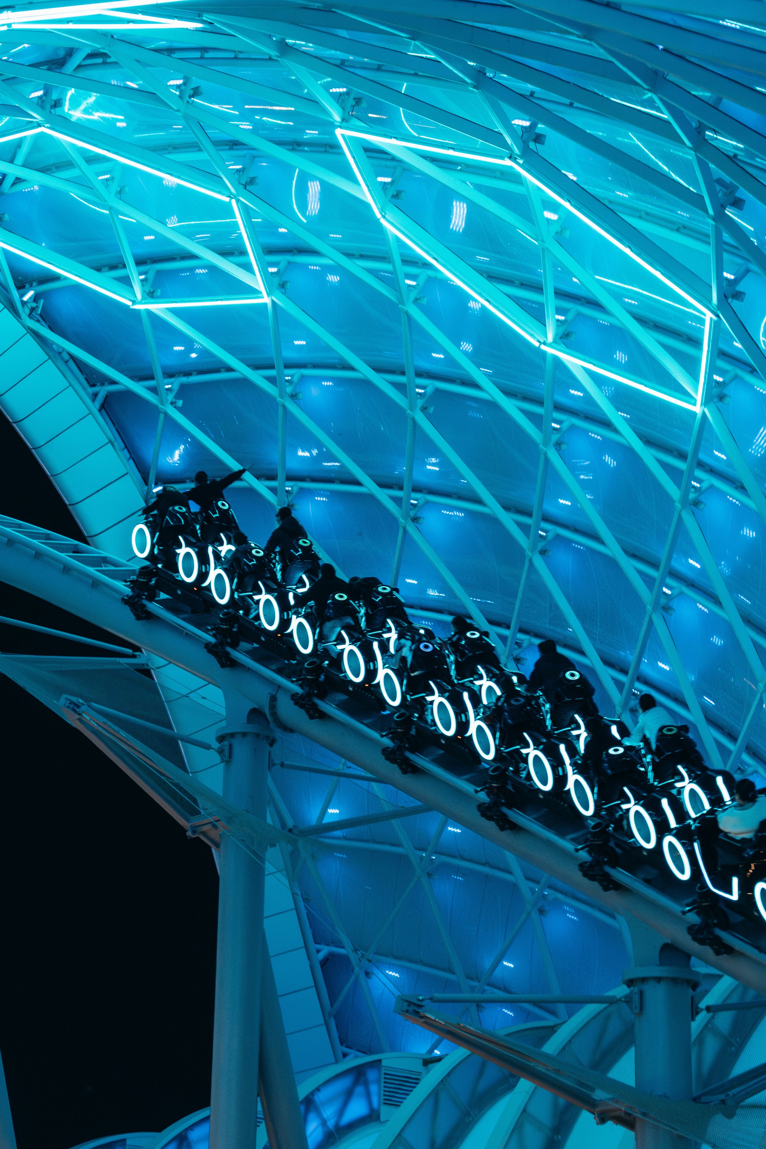 Nighttime view of a roller coaster with riders, illuminated with blue neon lights, beneath a covered structure with geometric patterns.