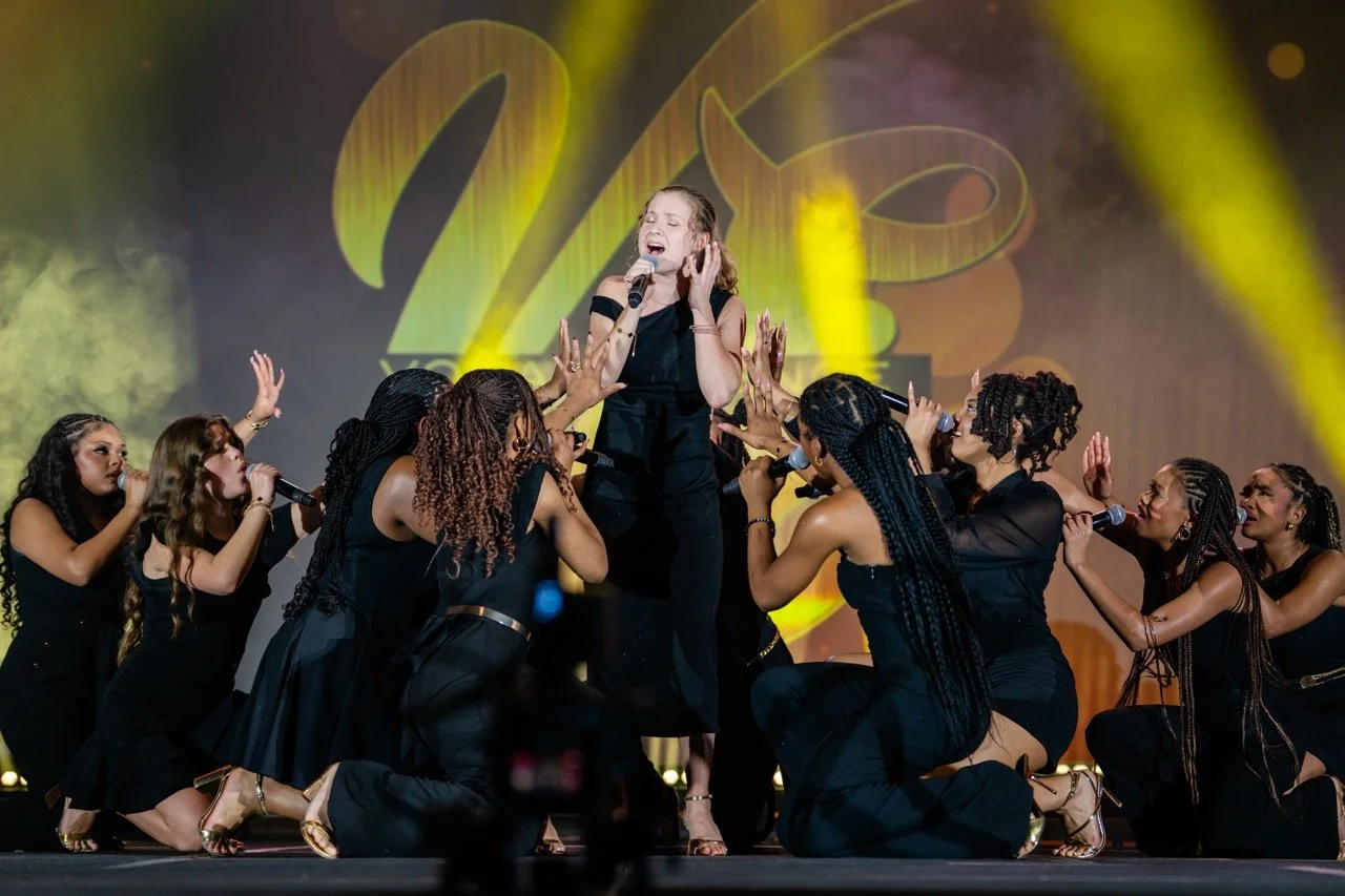 A woman singing on stage surrounded by a group of women, with a large illuminated number 20 in the background.