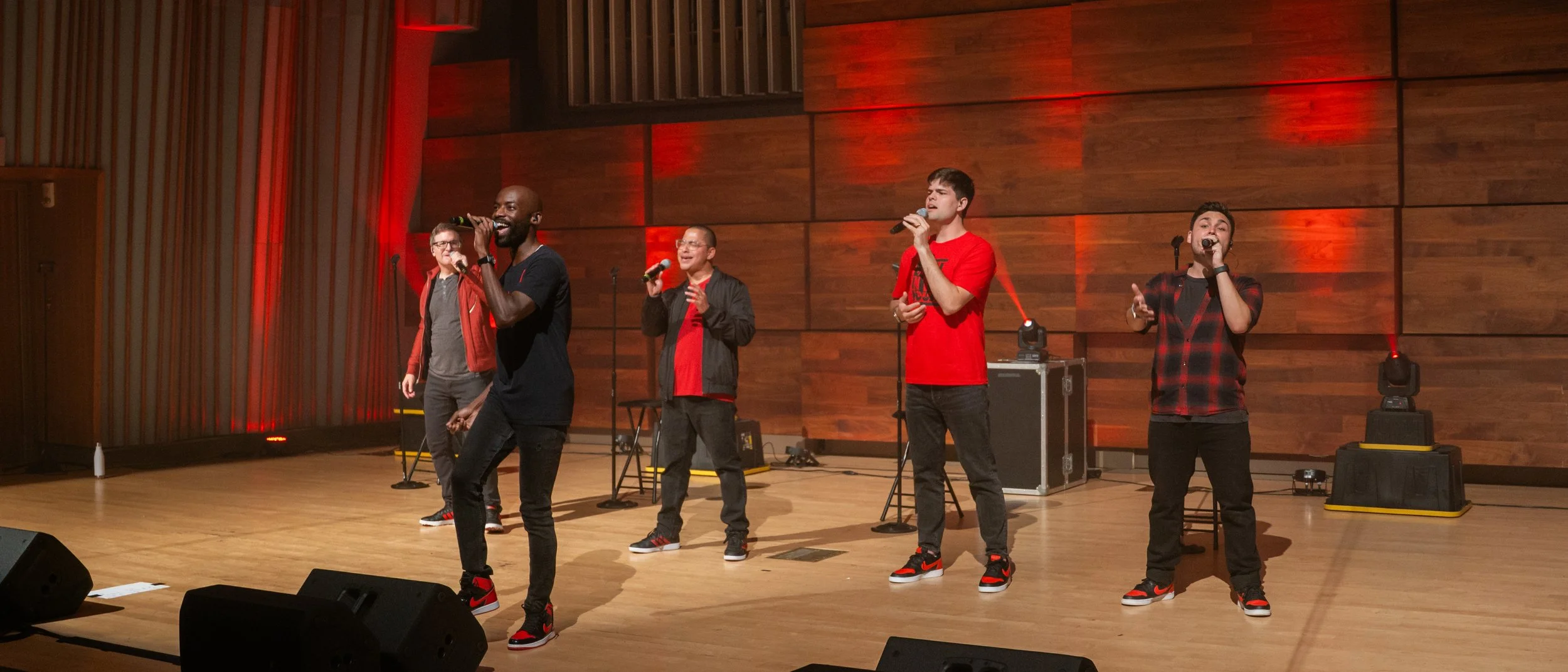 Five male singers performing on stage in a music venue with wooden wall backdrop, red lighting accents, microphones, and sound equipment.