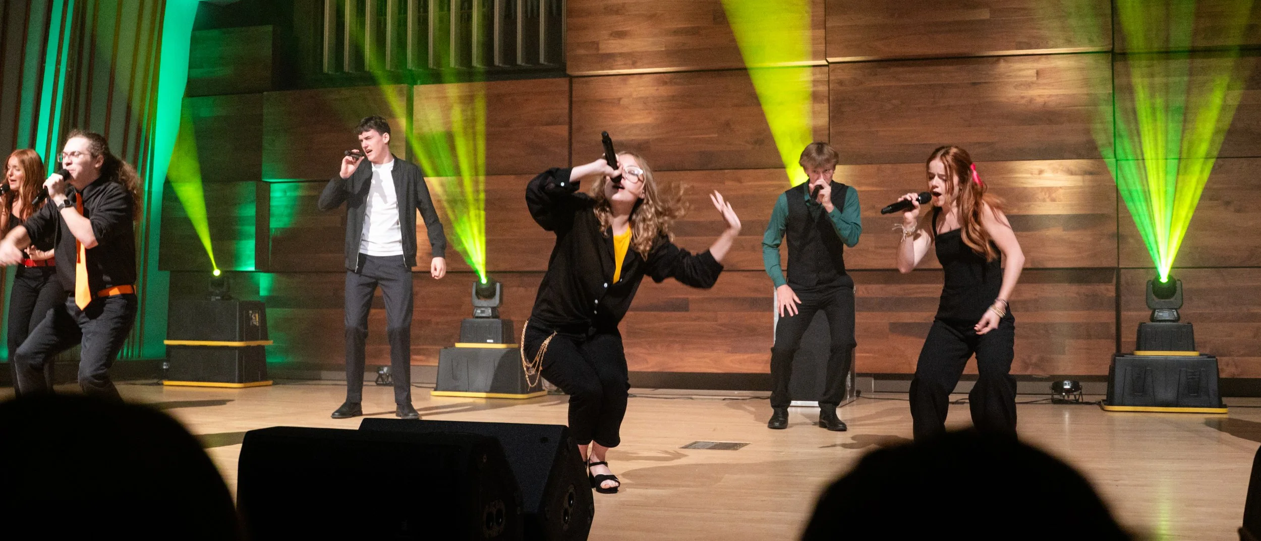 A group of six young performers, three women and three men, singing and dancing on stage with wooden panel background and green stage lights.