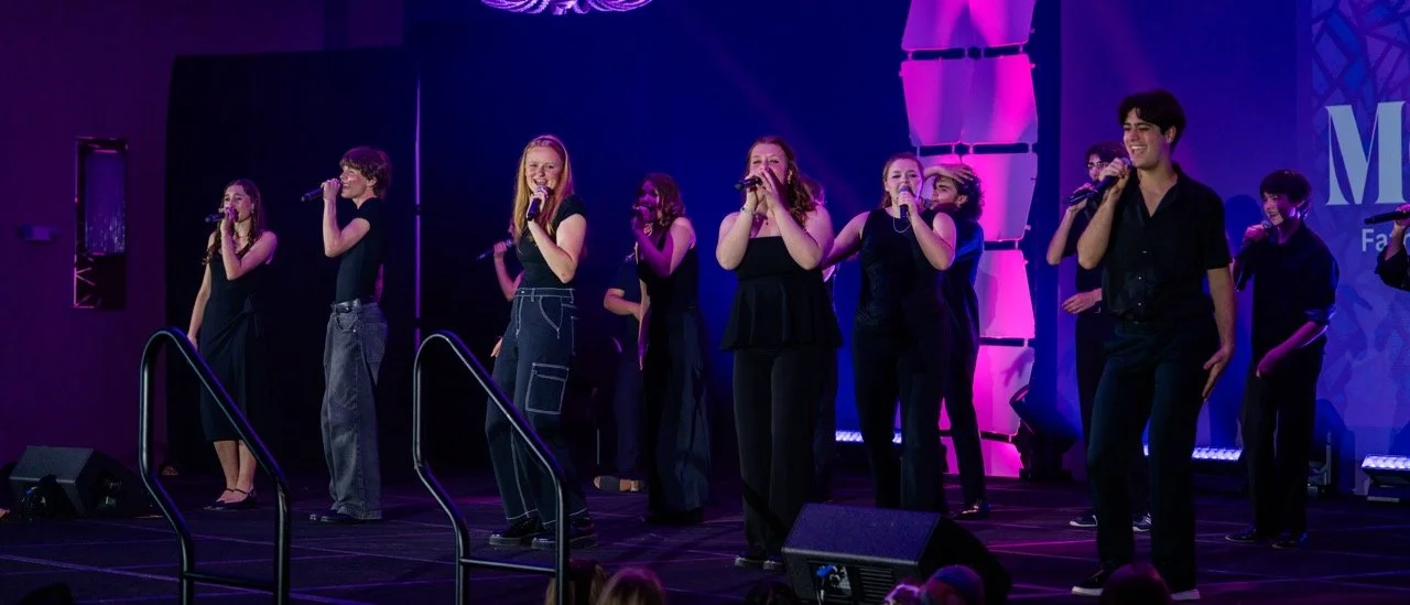 Group of young performers on stage singing into microphones during a live performance with purple and blue lighting.