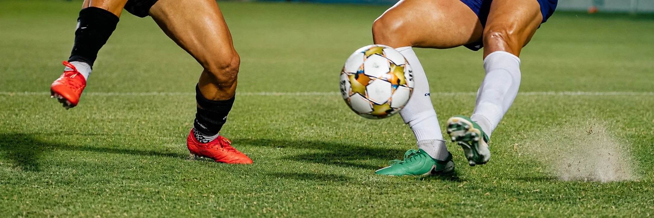 Two soccer players are battling for control of the ball on a grassy field, with one player wearing red shoes and black socks, and the other wearing green shoes and white socks.