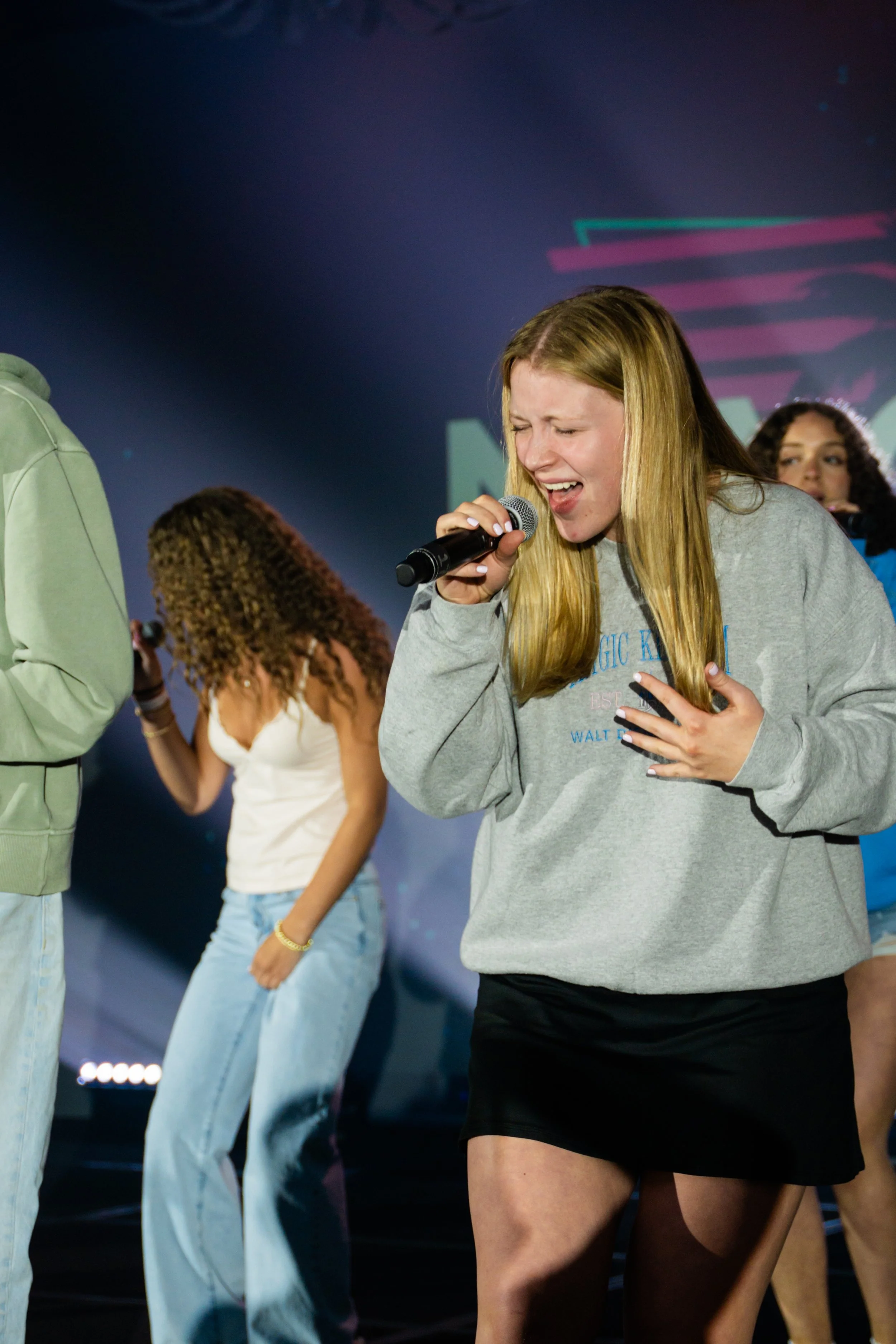 A group of young people singing and dancing on stage with a woman in the foreground holding a microphone, expressing emotion.