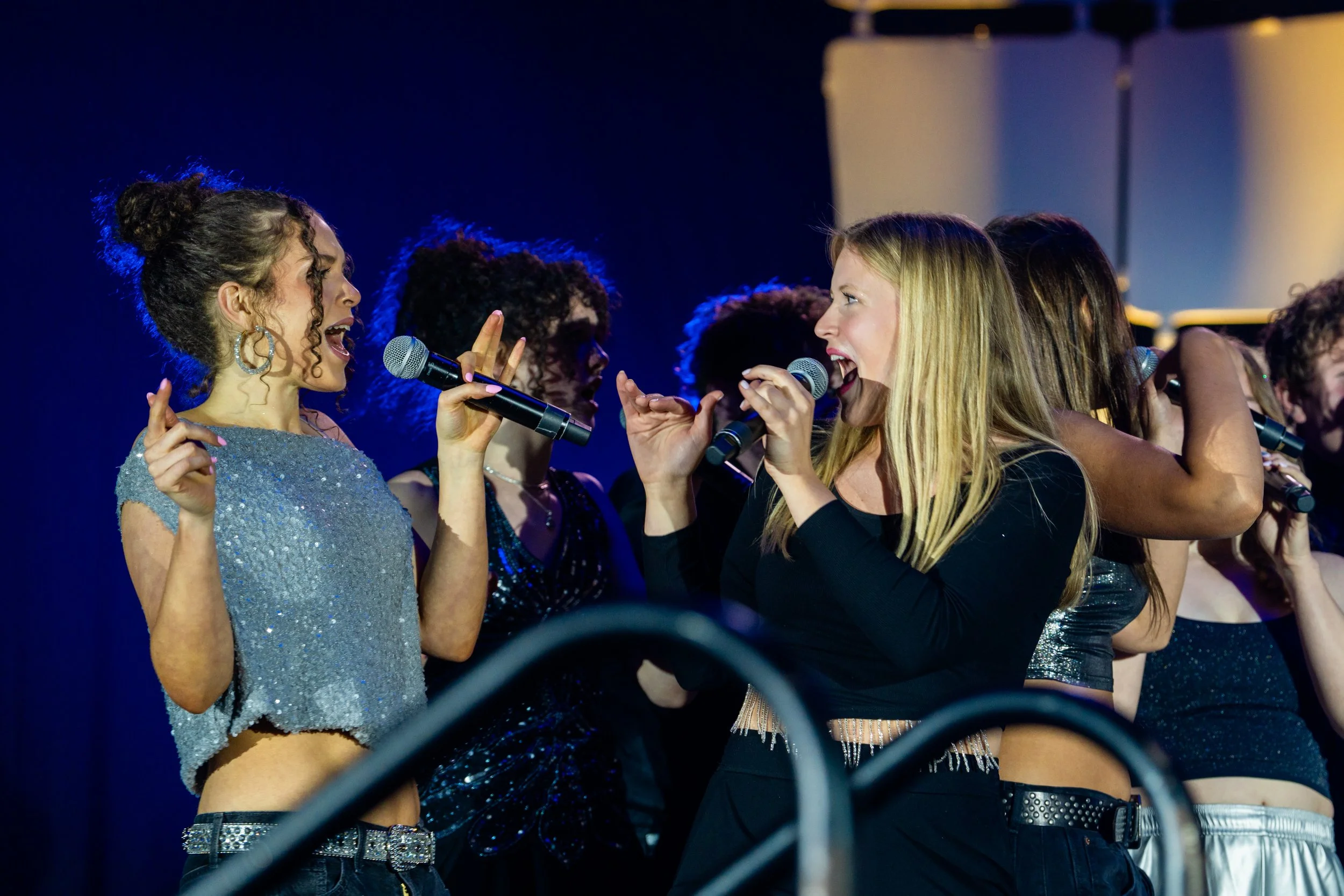Two women are facing each other, singing into microphones at a lively event, with a group of women in the background also holding microphones and singing.