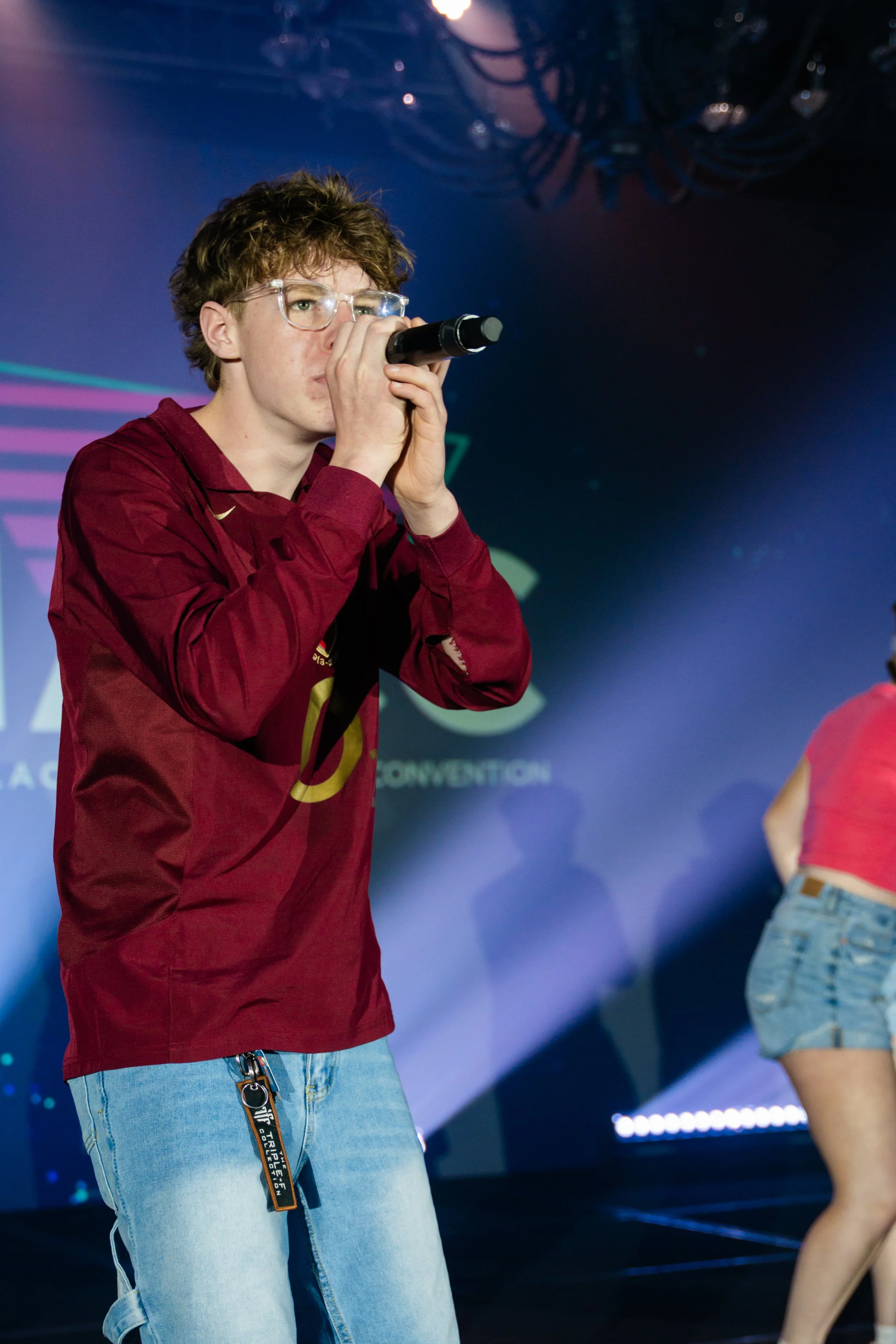 A person with curly brown hair, glasses, wearing a maroon jacket and light blue jeans, holding a microphone and speaking on stage. Part of another person in a pink top and denim shorts is visible to the right.
